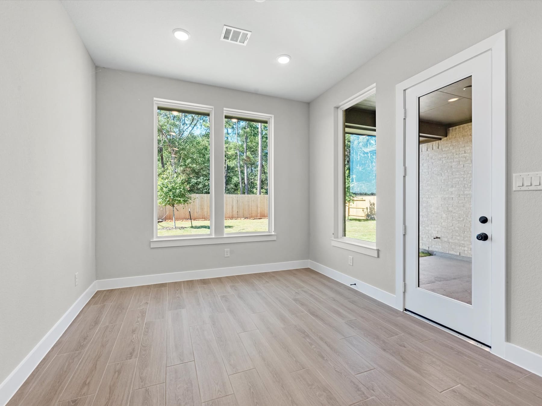 A bright and spacious room with large windows overlooking a lush, green outdoor area, featuring a hardwood floor and a white door leading to another part of the space.