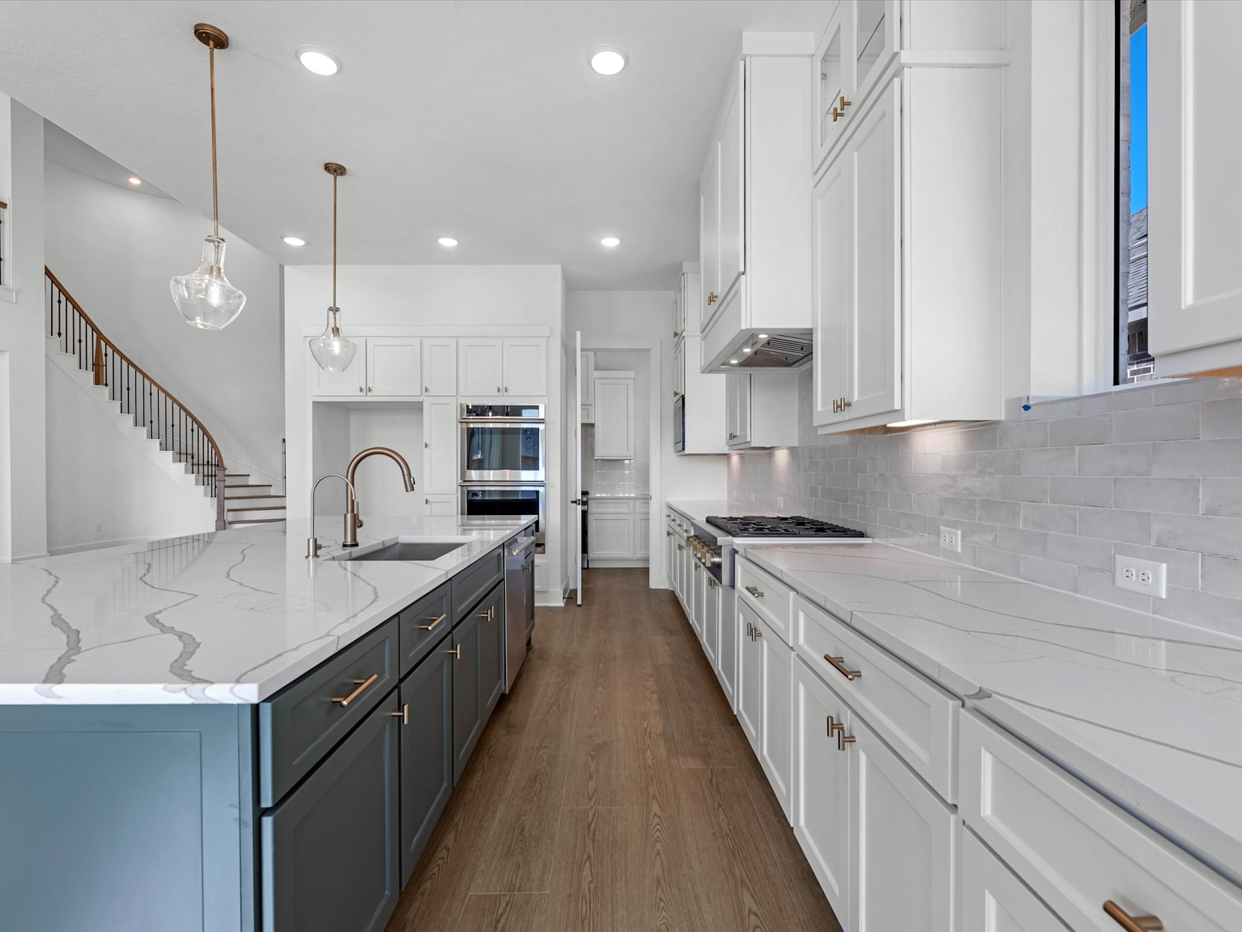 A modern, open-concept kitchen with white cabinets, dark countertops, and hardwood flooring, featuring pendant lighting and a staircase visible in the background.