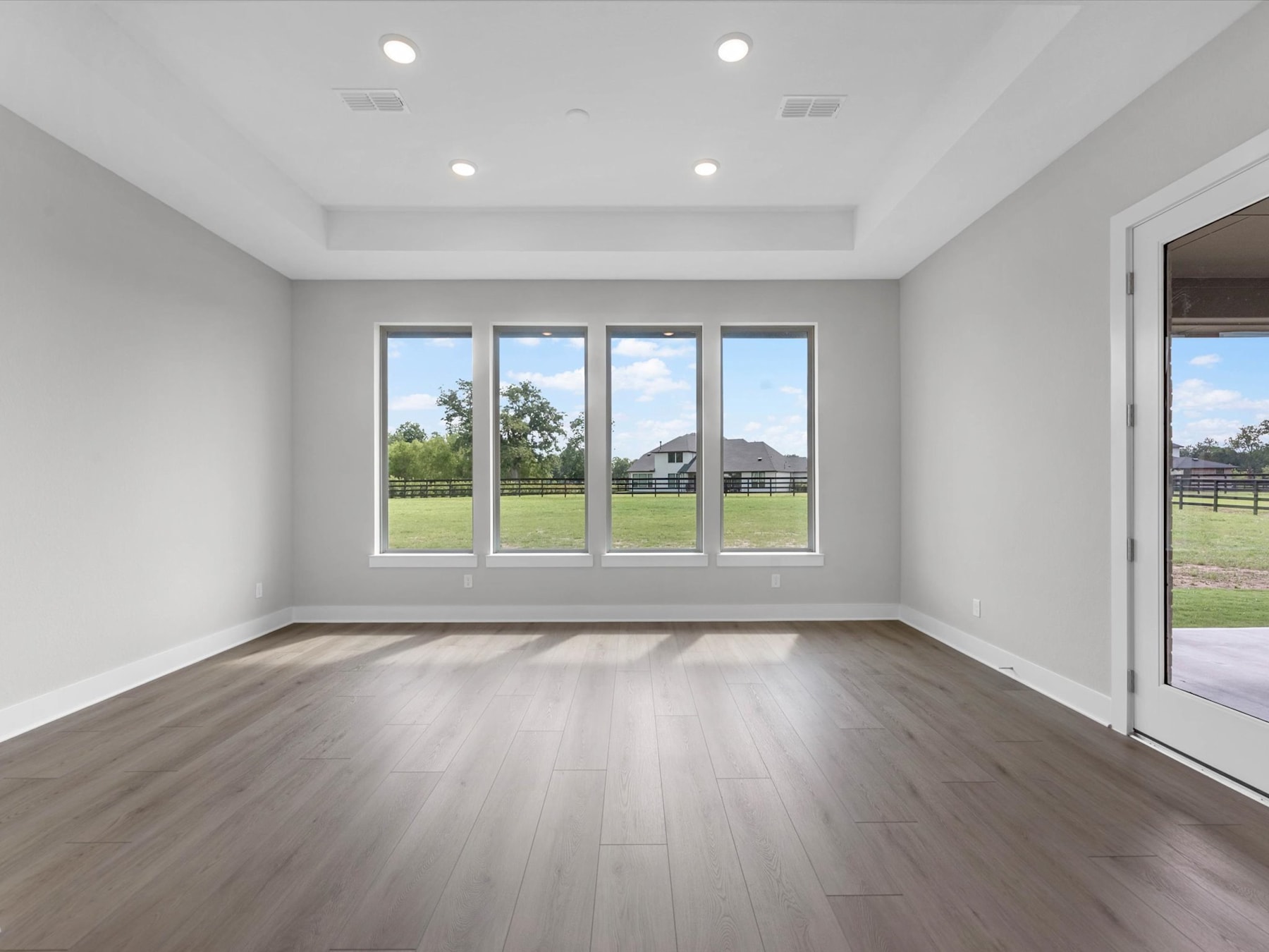 A spacious, well-lit room with large windows overlooking a grassy outdoor area, featuring a hardwood floor and recessed lighting.