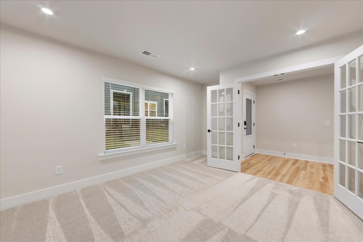 A bright and spacious room with hardwood floors, white walls, and a large window providing natural light. The room appears to be a living or bedroom space with a door leading to another area.