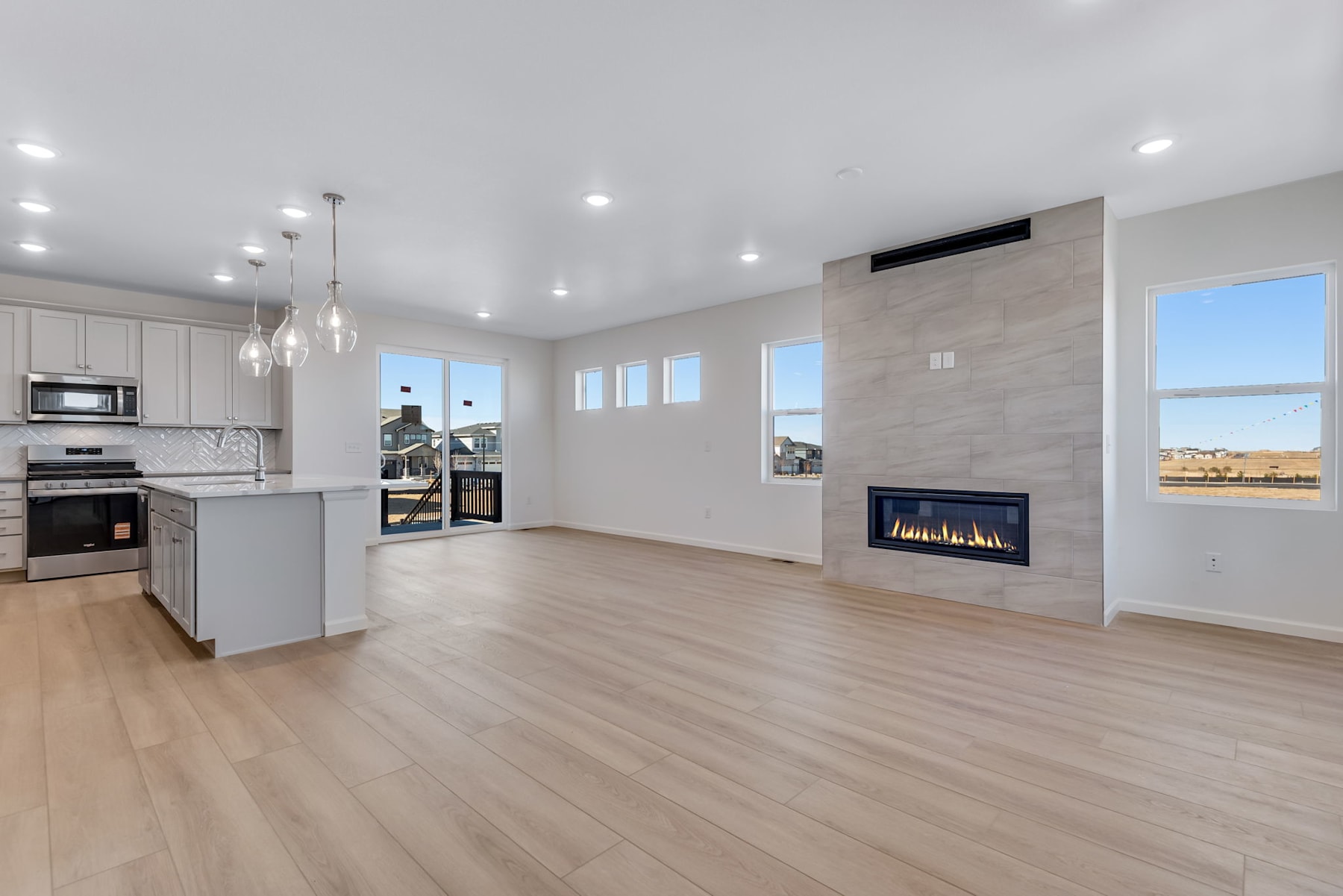 A modern, open-concept kitchen and living room with light-colored hardwood floors, white cabinets, stainless steel appliances, and a fireplace against a white wall.