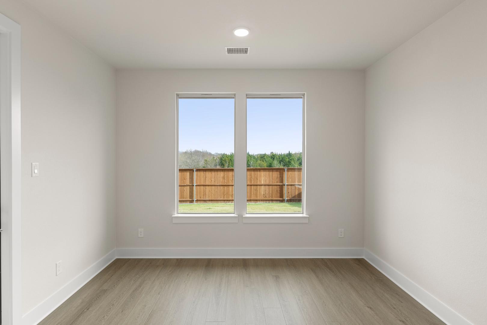 A bright, empty room with large windows overlooking a wooden fence and greenery in the background.