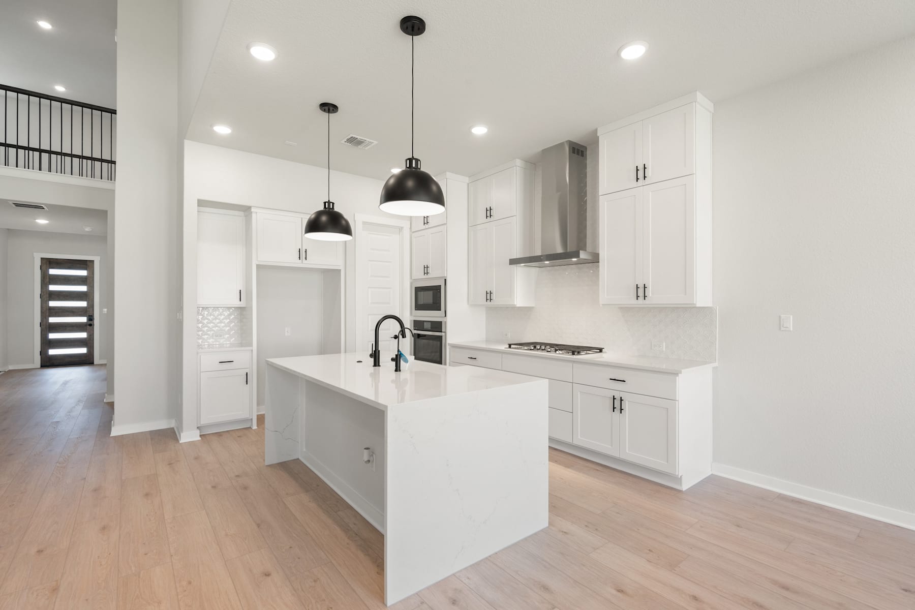A modern, minimalist kitchen with white cabinets, a central island, and pendant lighting fixtures, set against a backdrop of light-colored hardwood floors and walls.