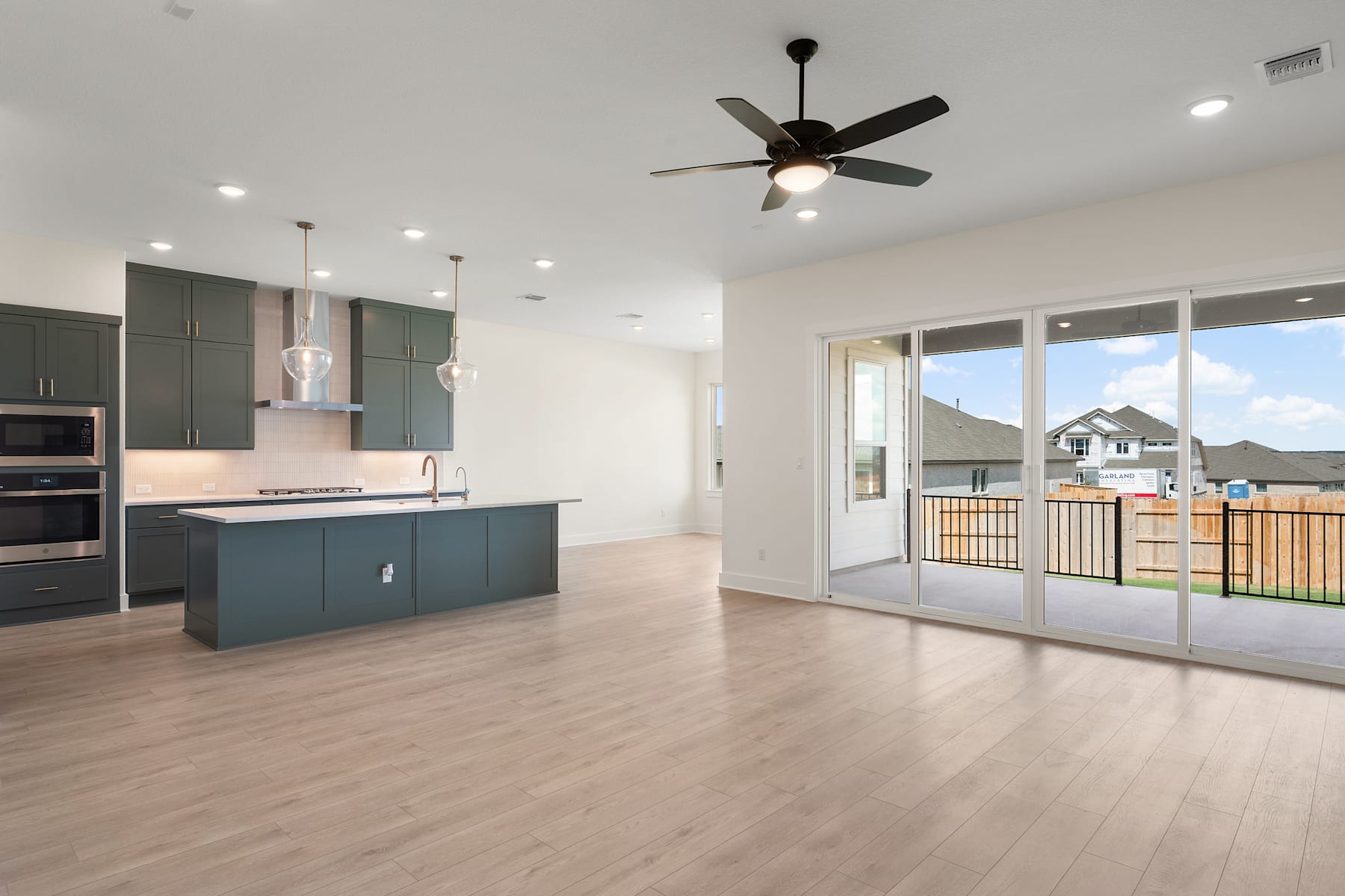 A modern, open-concept kitchen and living room with sleek gray cabinets, a ceiling fan, and large windows overlooking a backyard with a wooden fence.