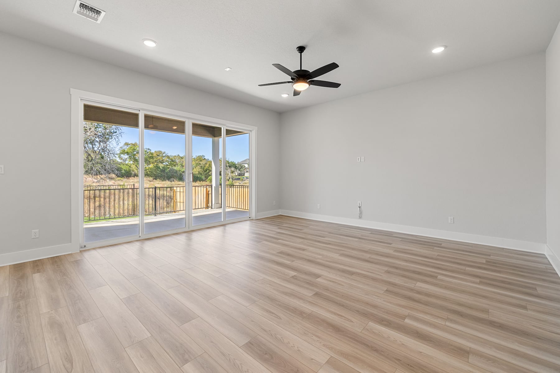 A spacious, well-lit room with a ceiling fan, hardwood floors, and large windows overlooking a backyard.