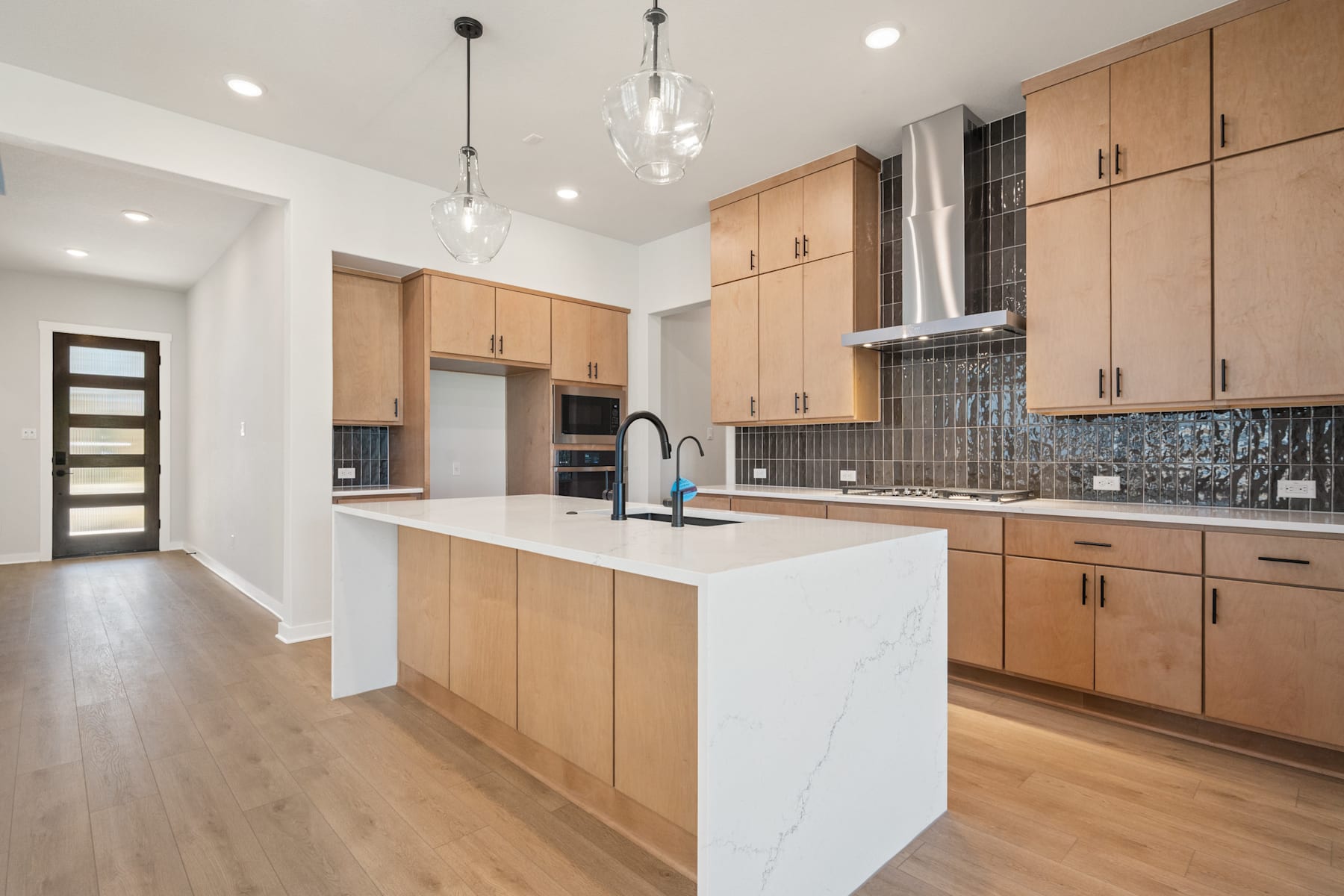 A modern, open-concept kitchen with light wood cabinets, a white countertop island, and pendant lighting fixtures, set against a backdrop of hardwood floors and a doorway leading to another room.