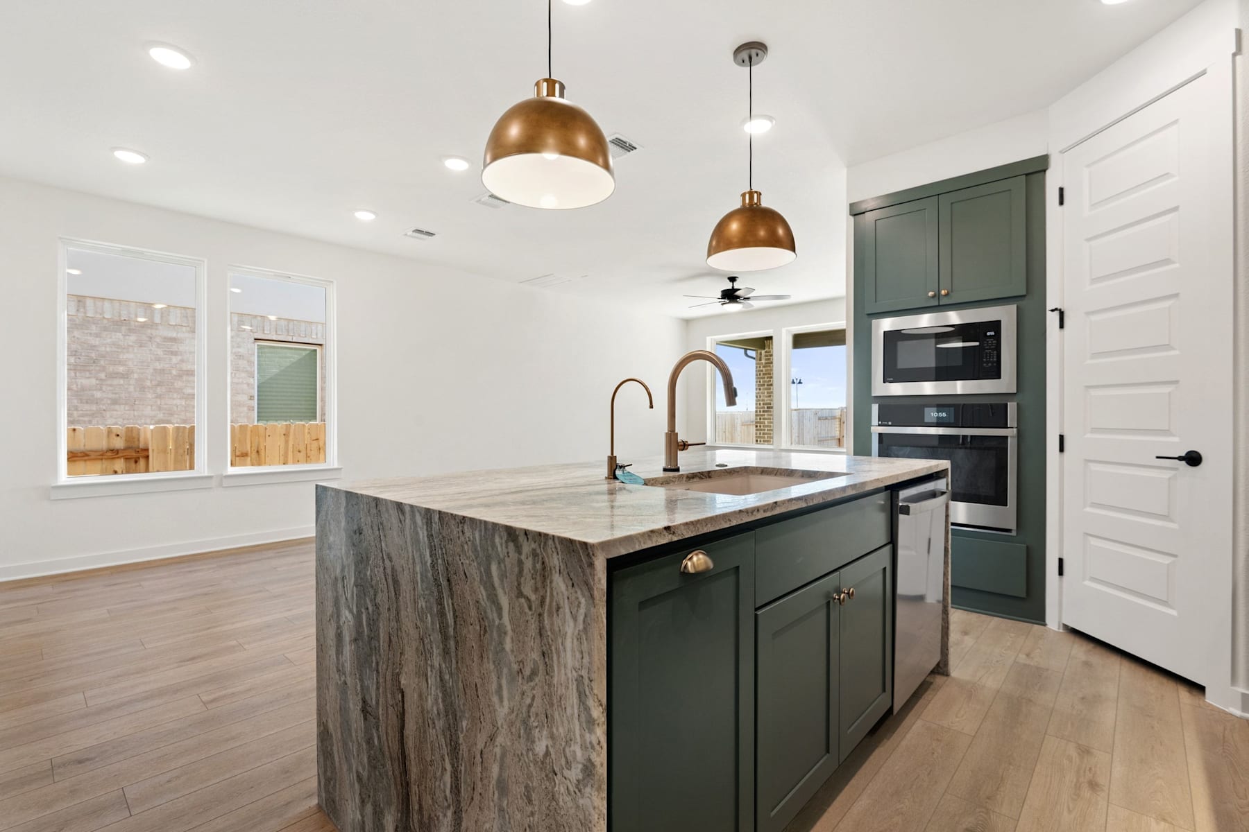 A modern and stylish kitchen with a large island, dark green cabinets, and pendant lights with a warm metallic finish, set against a bright and airy background.