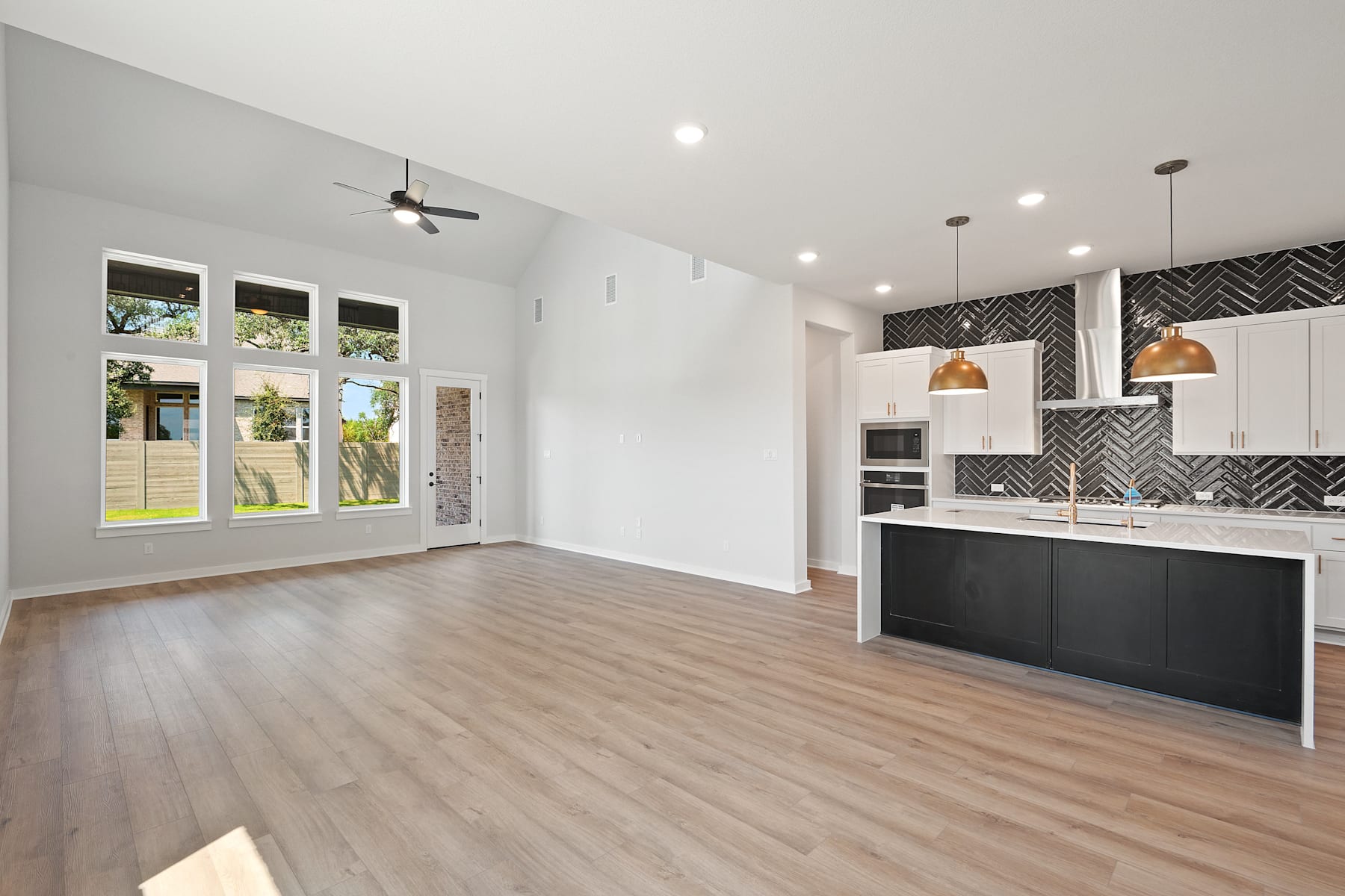 A modern, open-concept kitchen and living room with white walls, dark cabinets, and hardwood floors, featuring large windows that allow natural light to flood the space.