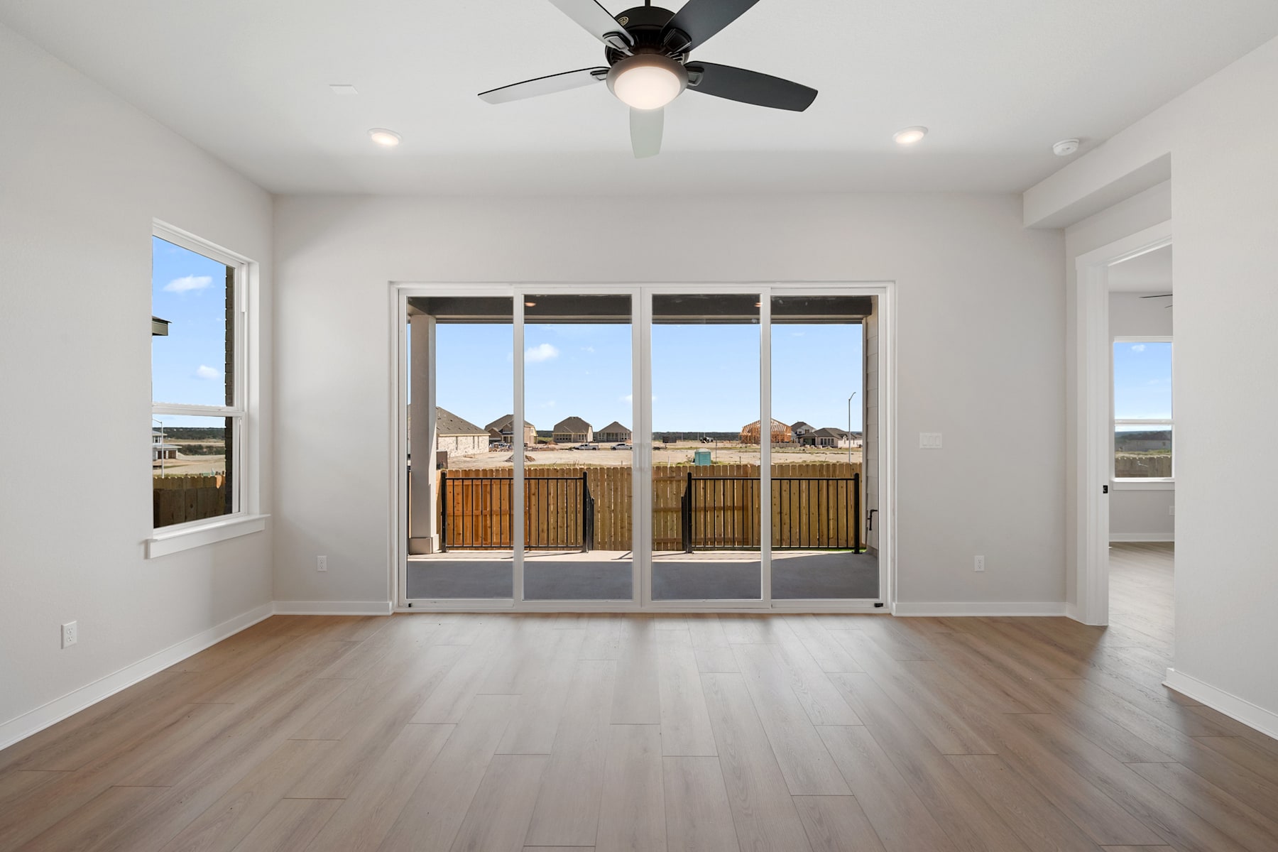 A spacious, well-lit room with a ceiling fan, wooden flooring, and large windows overlooking a residential neighborhood in the background.