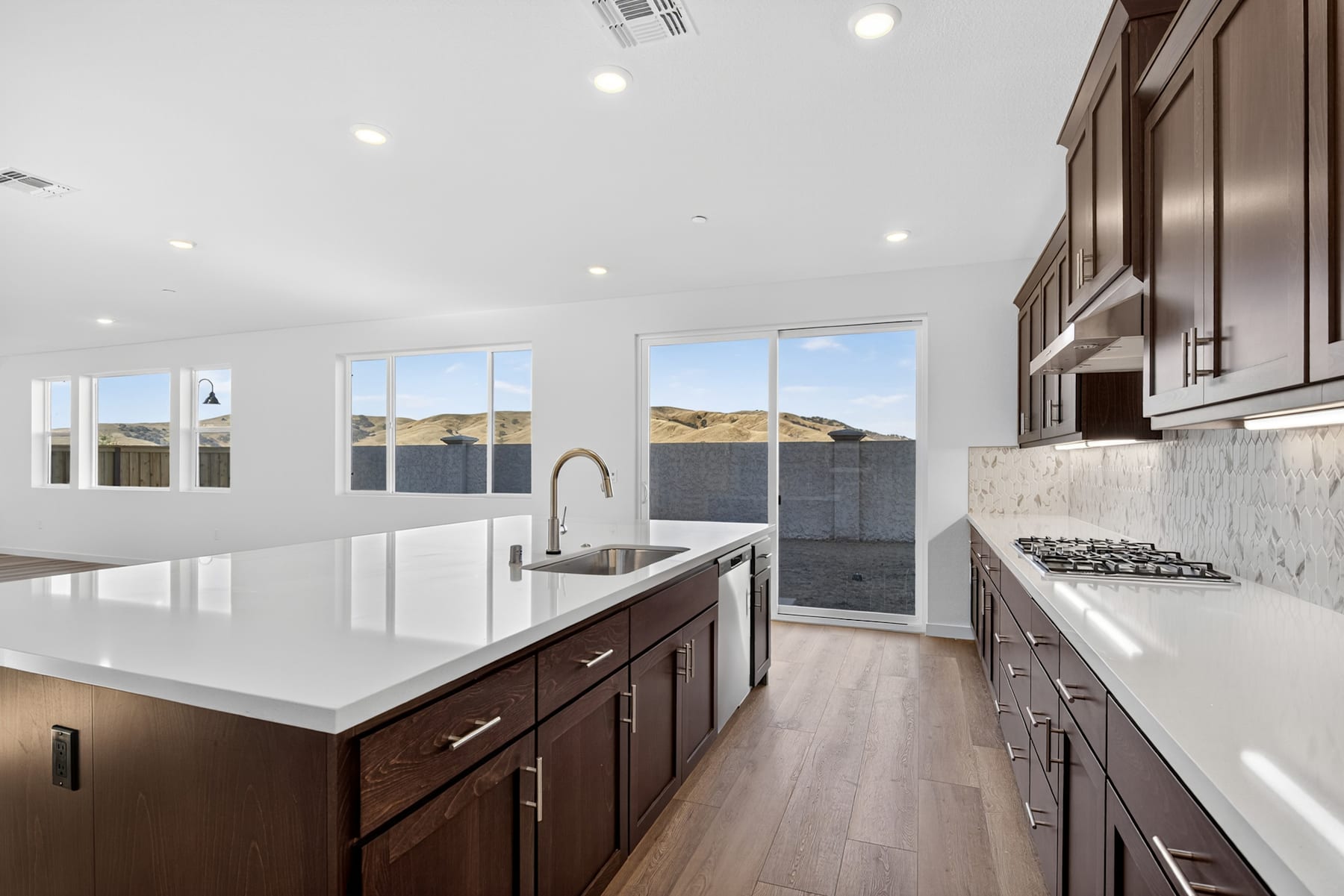 A modern, well-lit kitchen with dark wood cabinets, white countertops, and large windows overlooking a scenic outdoor landscape.