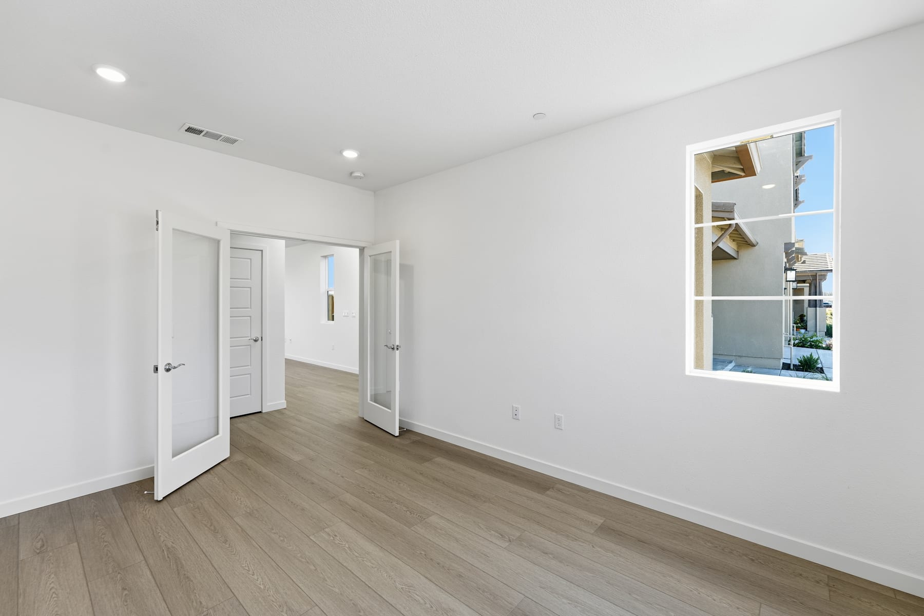 A spacious, well-lit room with white walls, a large window, and hardwood flooring. The room appears to be a bedroom or living space, with a door leading to another area.