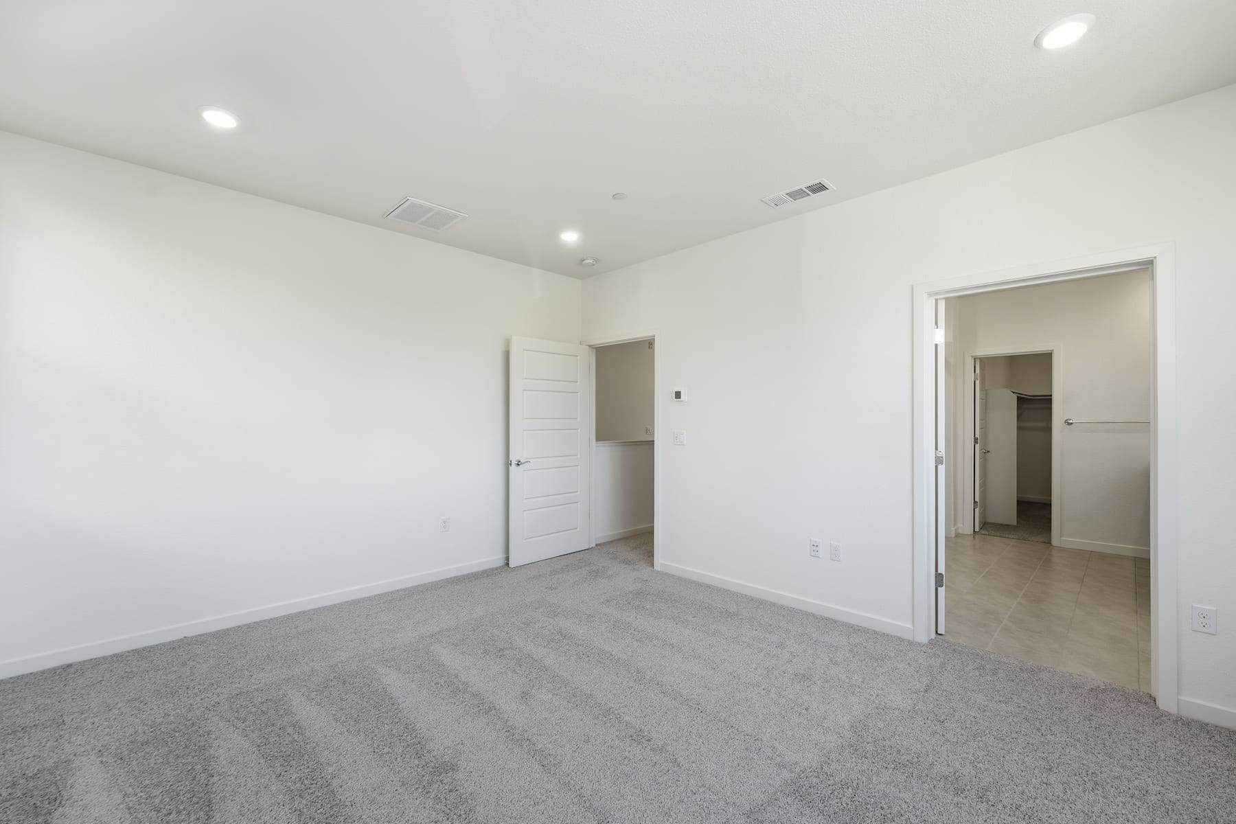 A spacious, minimalist bedroom with white walls, a gray carpeted floor, and a closed door leading to another room.