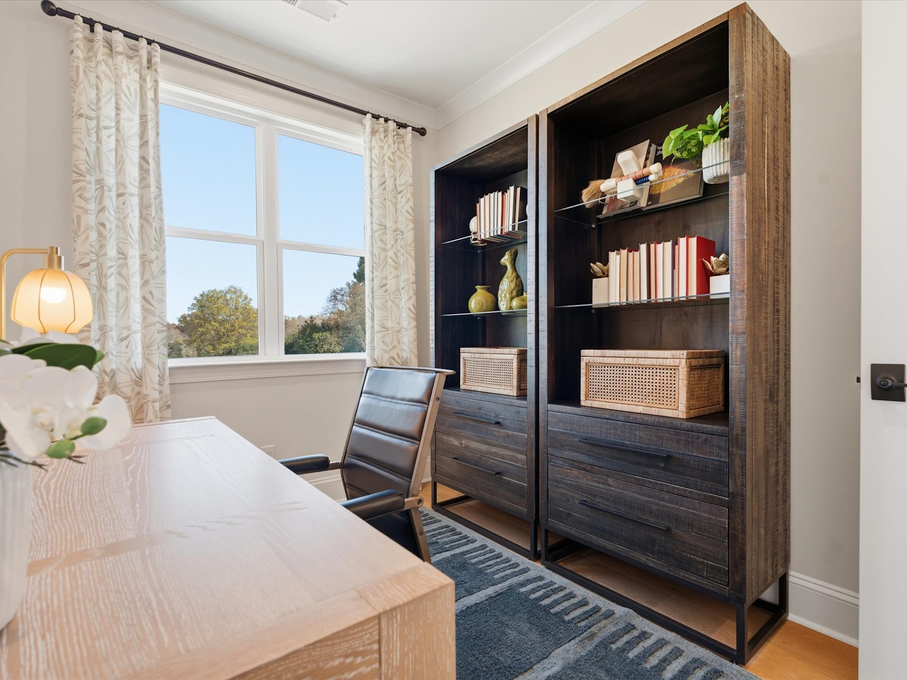 A cozy and well-organized home office space with a wooden desk, shelving unit, and decorative elements, set against a backdrop of a large window overlooking a scenic outdoor view.