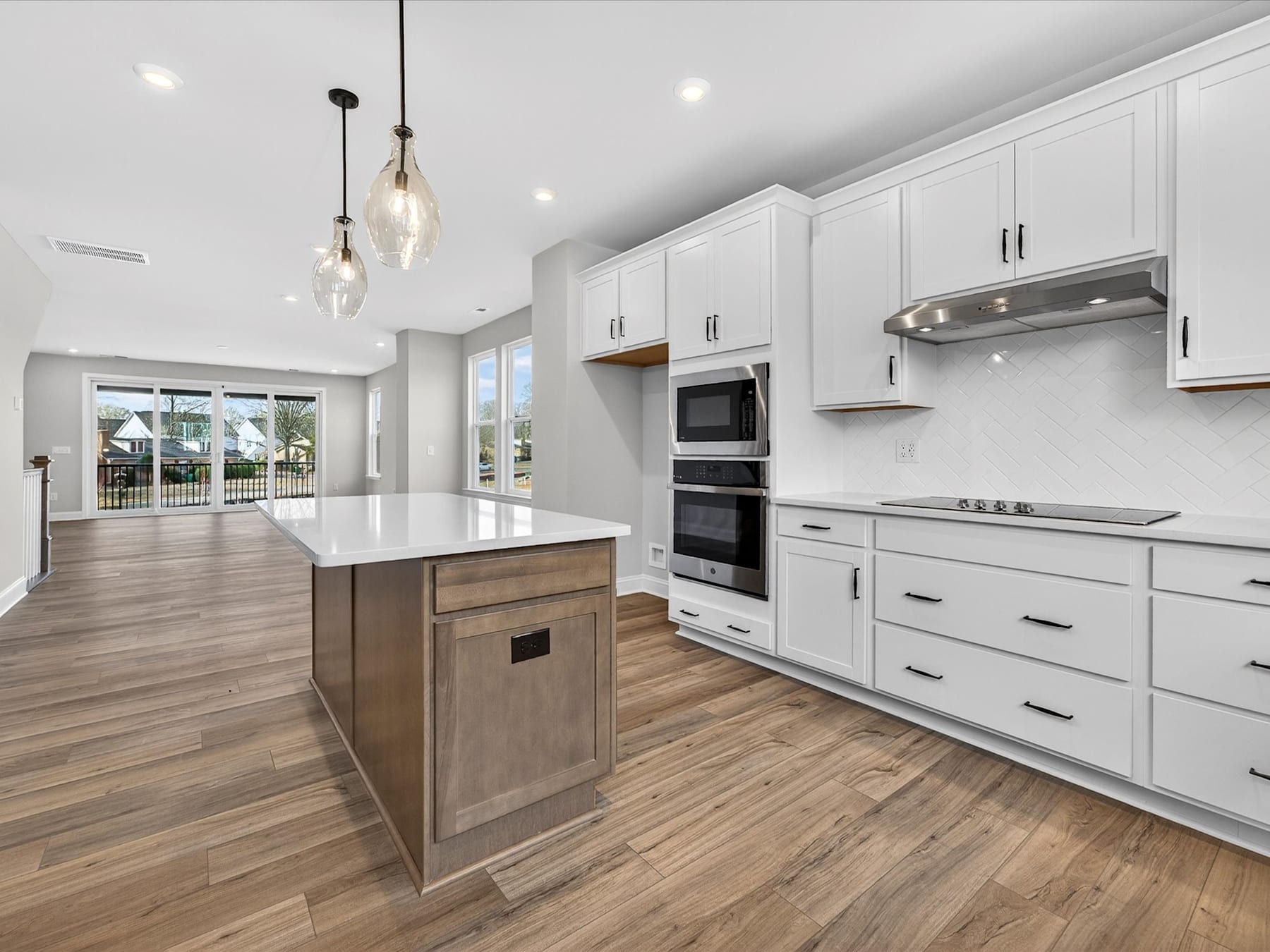 A modern, open-concept kitchen with white cabinets, a large island, and hardwood flooring, featuring pendant lighting and a view of the exterior through a sliding glass door.