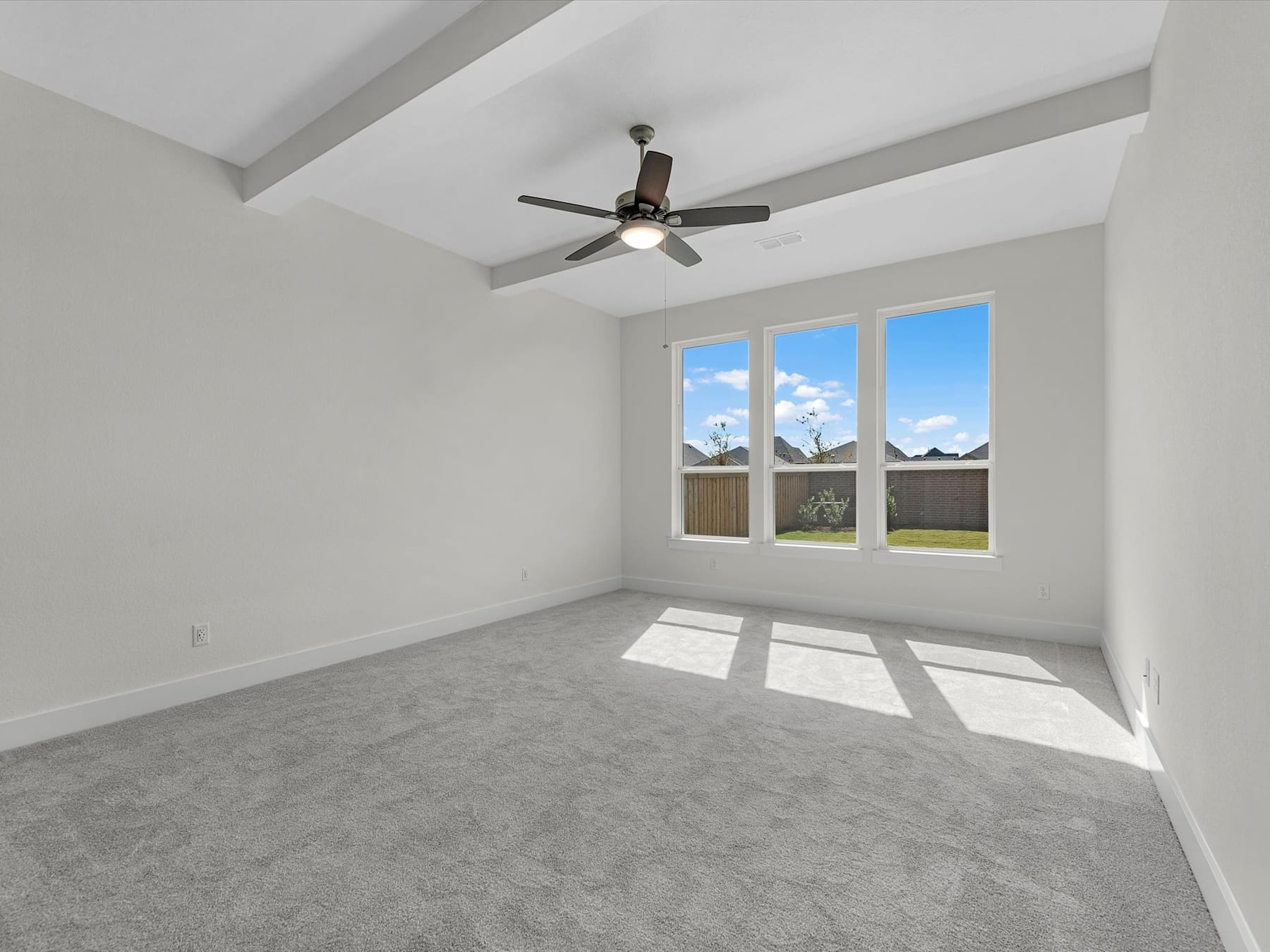 A spacious, well-lit room with a ceiling fan and large windows overlooking a backyard.