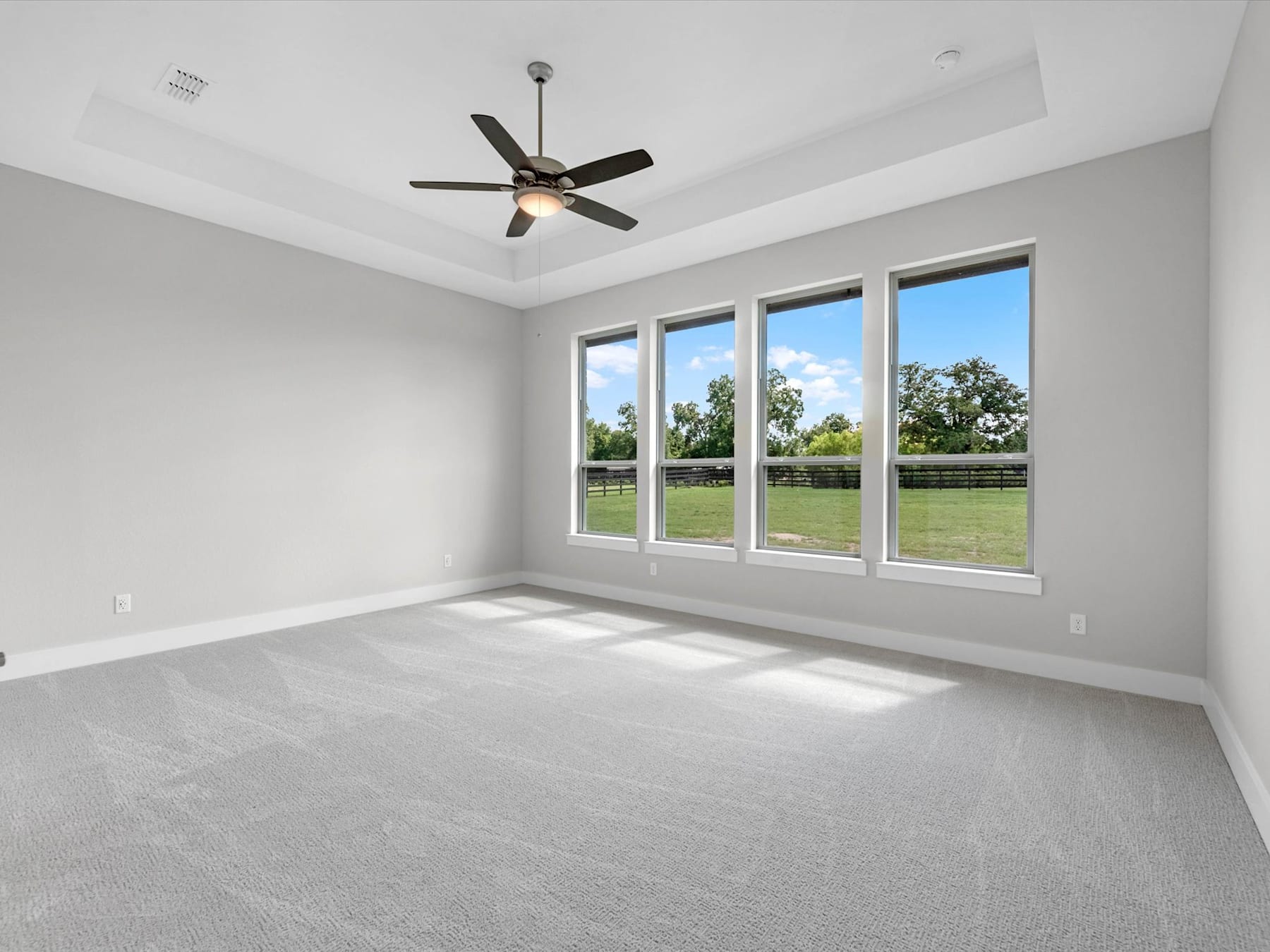 A spacious, bright room with large windows overlooking a grassy outdoor area, featuring a ceiling fan and neutral-toned flooring.