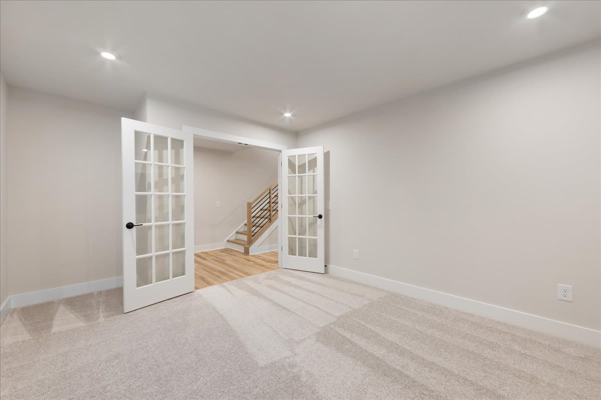 A bright and spacious room with white walls, hardwood floors, and a set of French doors leading to a staircase in the background.