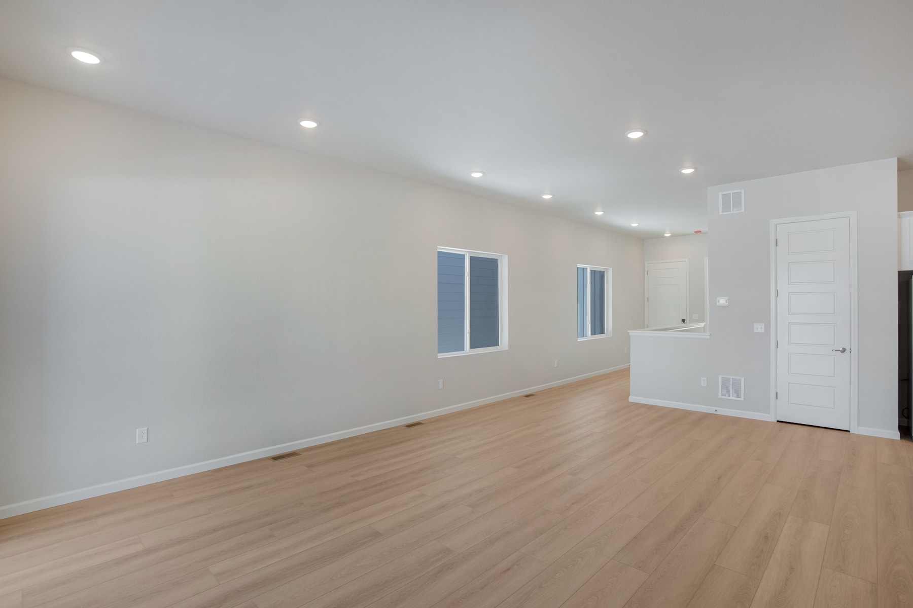 A spacious, well-lit room with a hardwood floor and white walls, featuring recessed lighting and a door leading to another area.