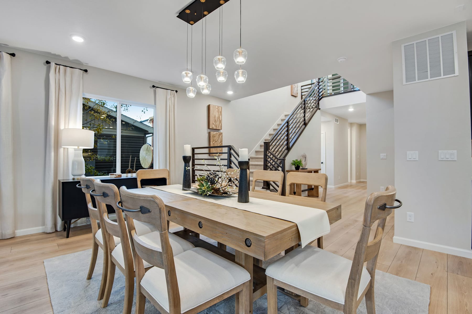 A modern and minimalist dining room with a wooden table, chairs, and a chandelier with hanging glass lights, set against a backdrop of a staircase and a view of the outdoors through a large window.