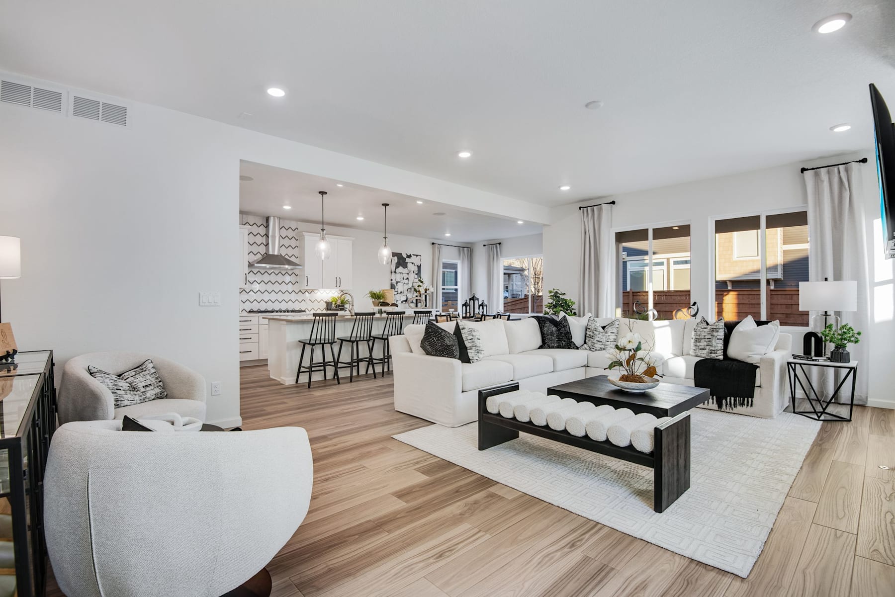 A modern and spacious open-concept living room with a white sofa, a black coffee table, and a kitchen visible in the background, all set against a backdrop of light-colored hardwood floors and white walls.