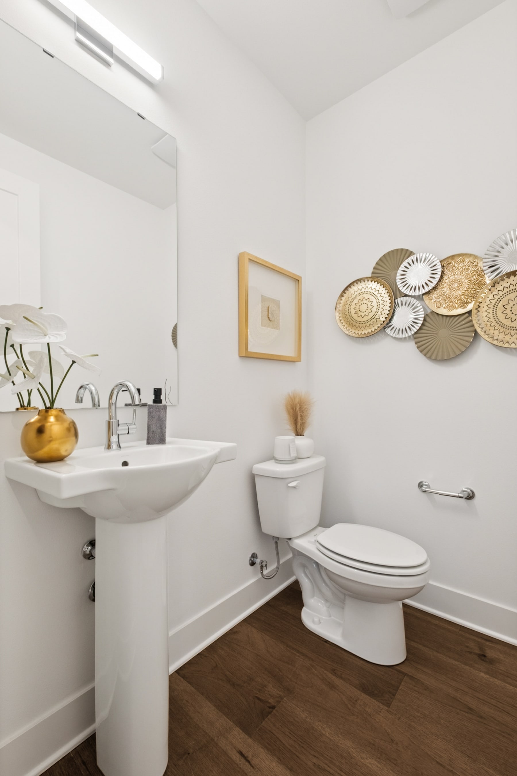 A modern and minimalist bathroom with a white pedestal sink, a toilet, and decorative wall hangings, set against a backdrop of white walls and a wooden floor.