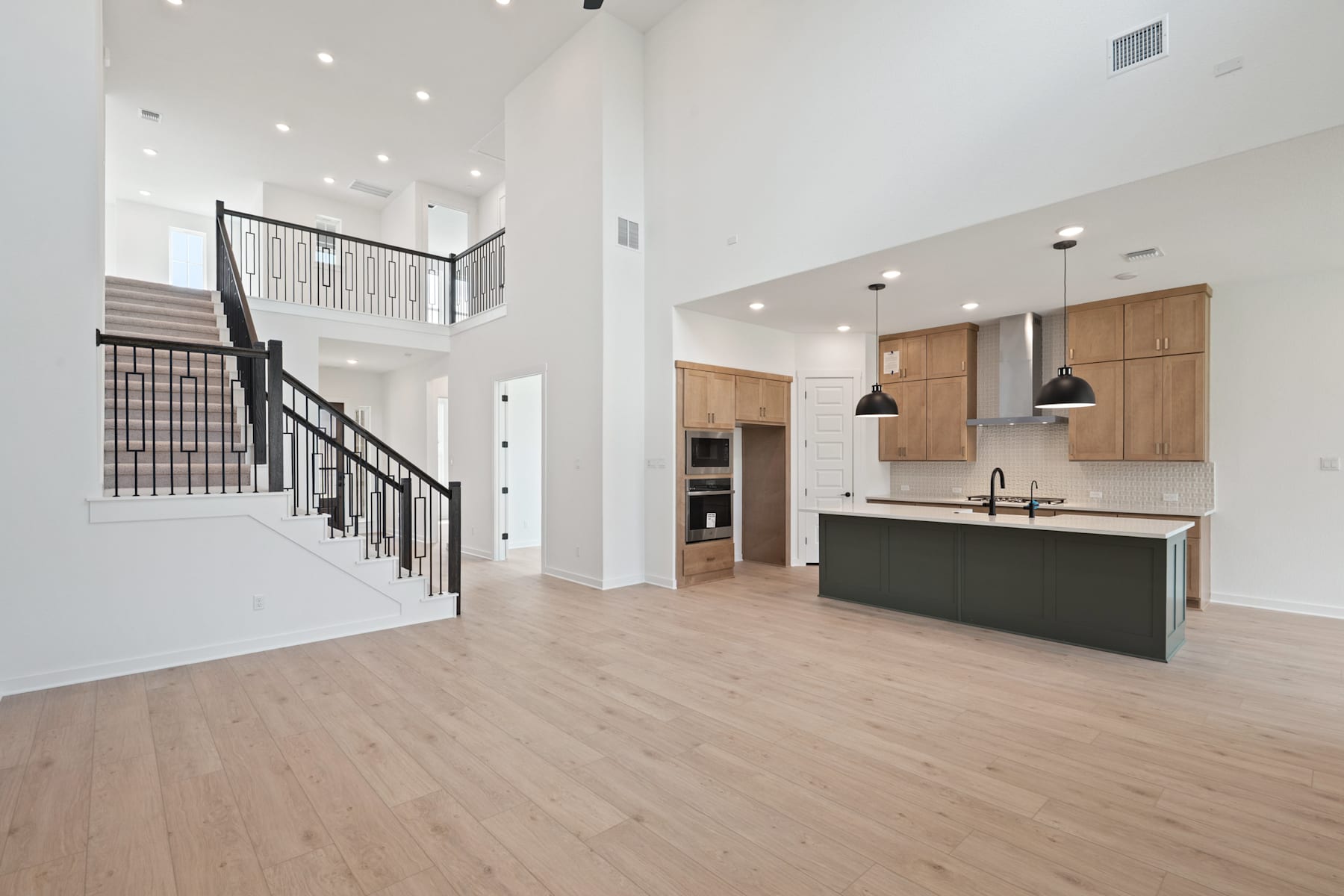 A spacious and modern open-concept living space with a kitchen, dining area, and staircase leading to an upper level. The room features high ceilings, hardwood floors, and a mix of light and dark wood tones in the cabinetry and furnishings.