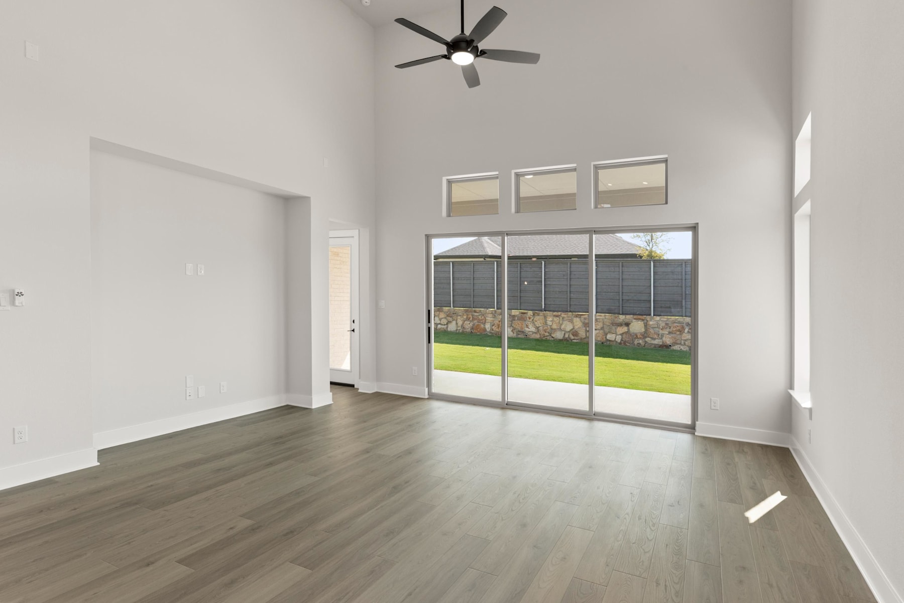 A spacious, well-lit room with a ceiling fan, hardwood floors, and large sliding glass doors leading to a grassy outdoor area.