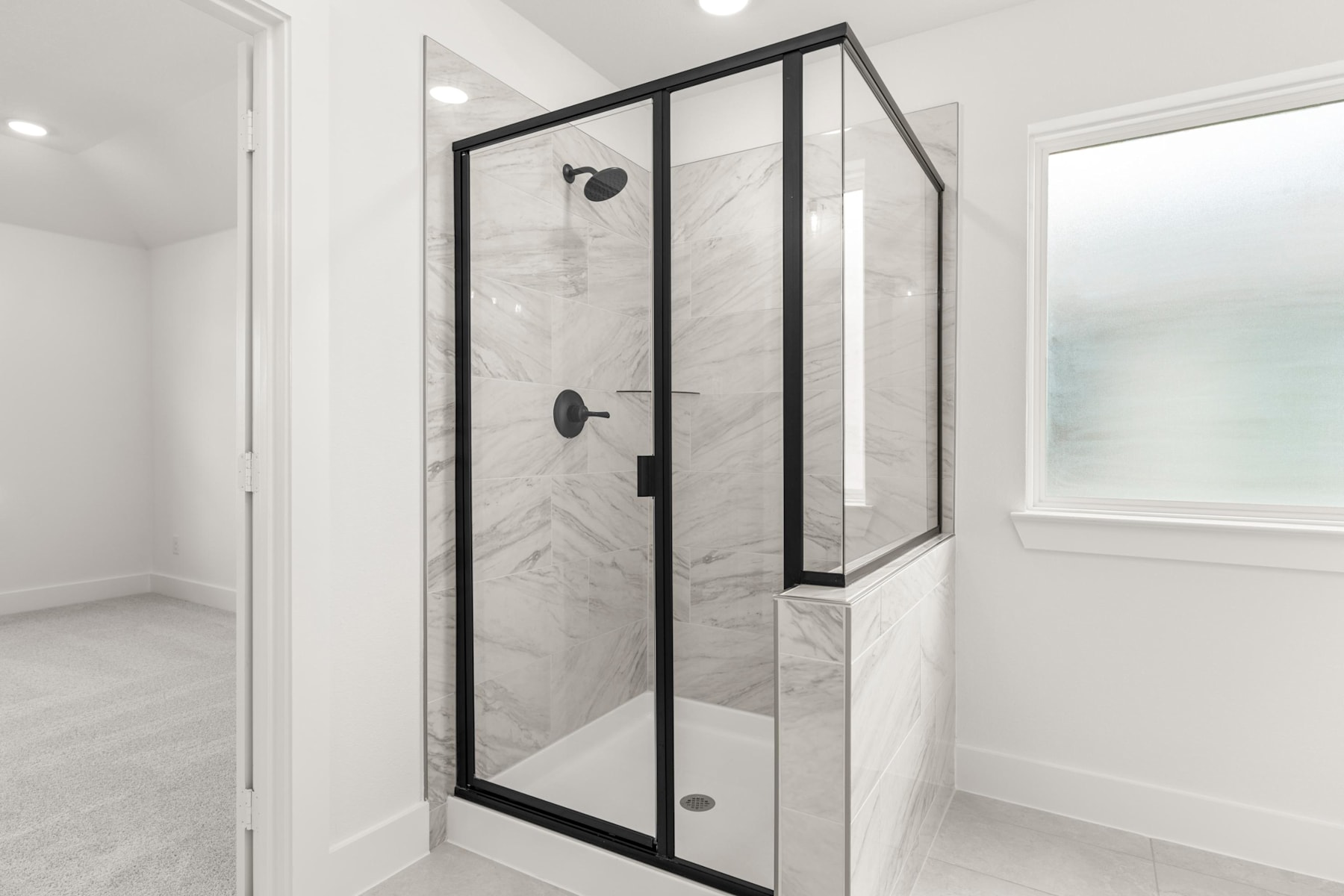 A modern, minimalist bathroom with a glass-enclosed shower stall featuring a black metal frame, surrounded by white walls and a tiled floor.