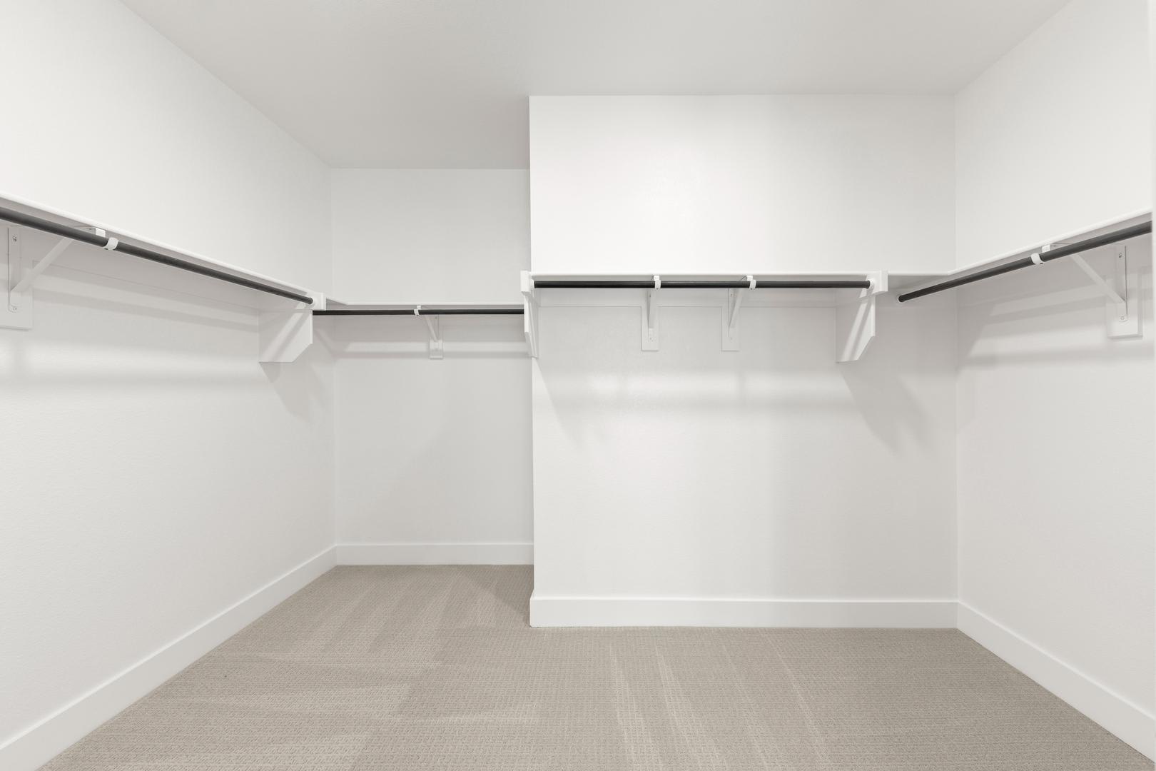 A spacious, well-lit walk-in closet with white walls, a wooden floor, and a series of metal hanging rods for storing clothes.