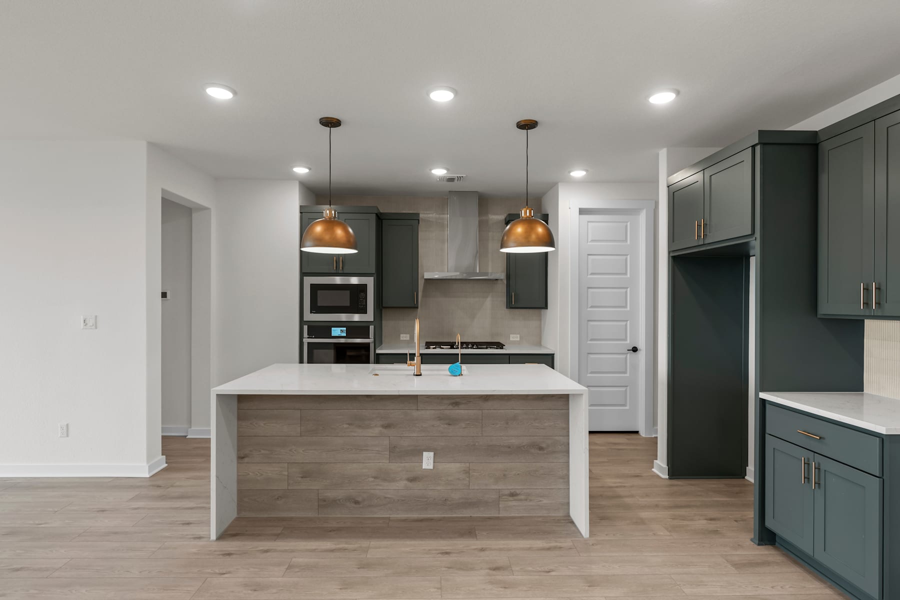 A modern and spacious kitchen with a central island, dark green cabinets, and pendant lighting fixtures, set against a backdrop of white walls and hardwood flooring.