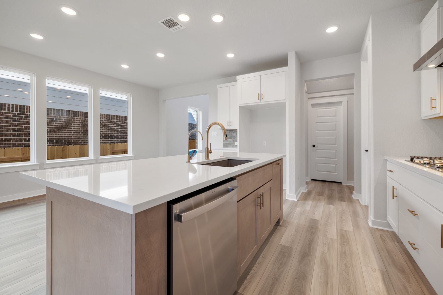A modern, open-concept kitchen with white countertops, light wood cabinets, and hardwood flooring, featuring a central island and recessed lighting.