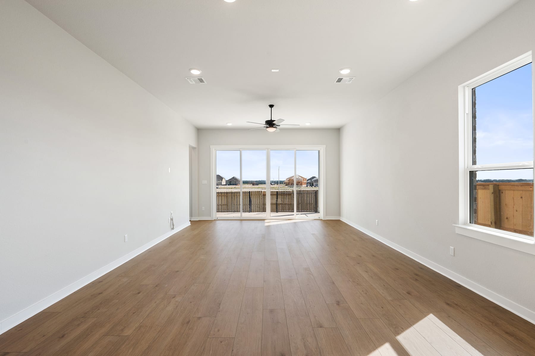 A spacious, well-lit room with hardwood floors, recessed lighting, and large windows overlooking a backyard.