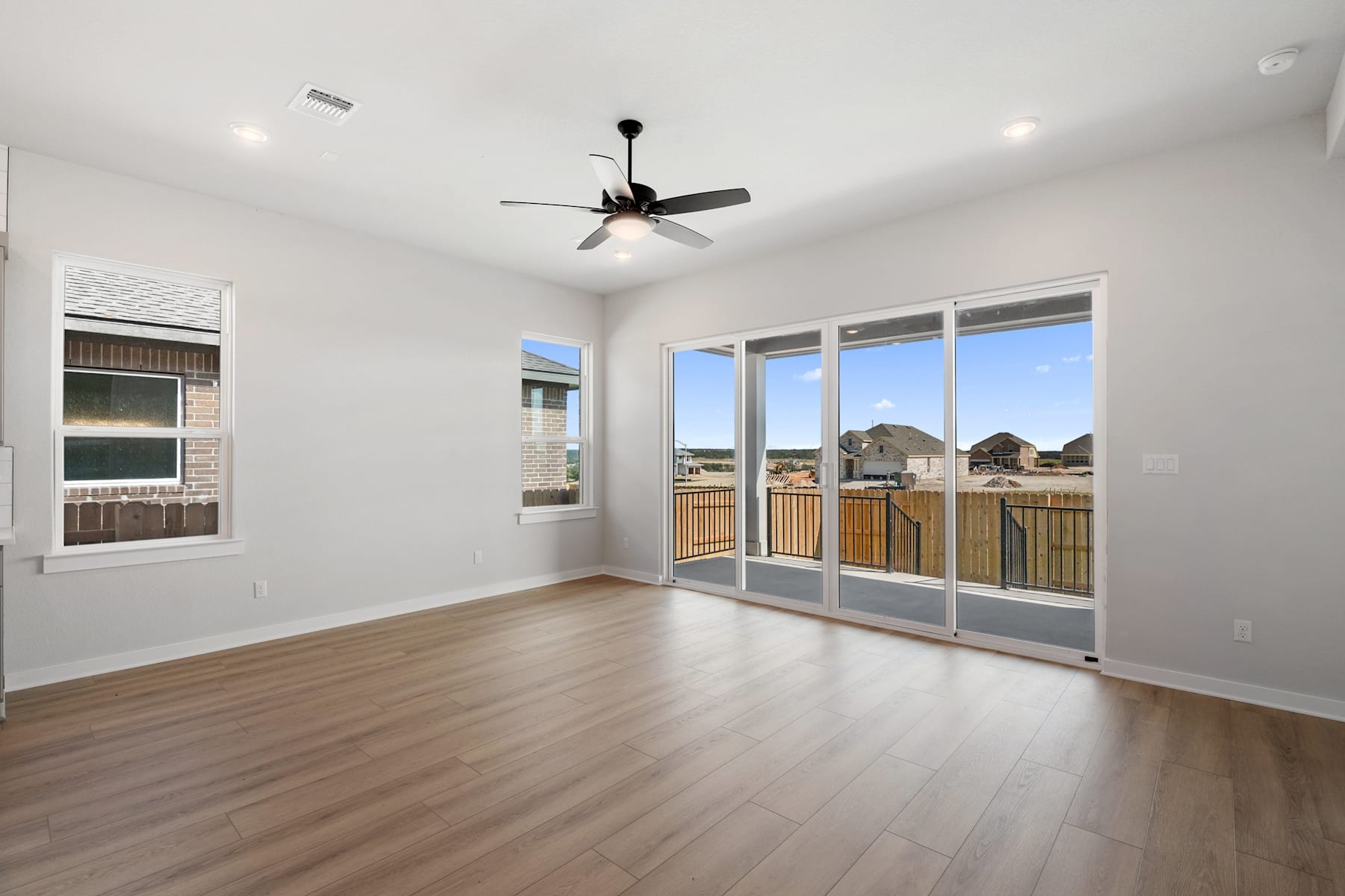 A spacious, well-lit room with a ceiling fan, hardwood floors, and large windows overlooking a residential neighborhood in the background.