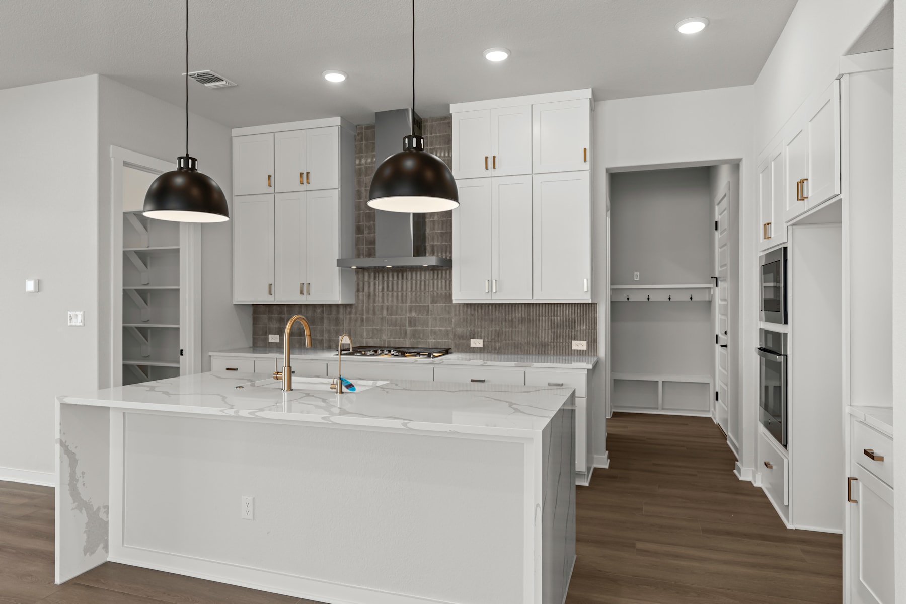 A modern, minimalist kitchen with white cabinets, a central island, and pendant lighting fixtures, set against a backdrop of hardwood floors and a doorway leading to another room.