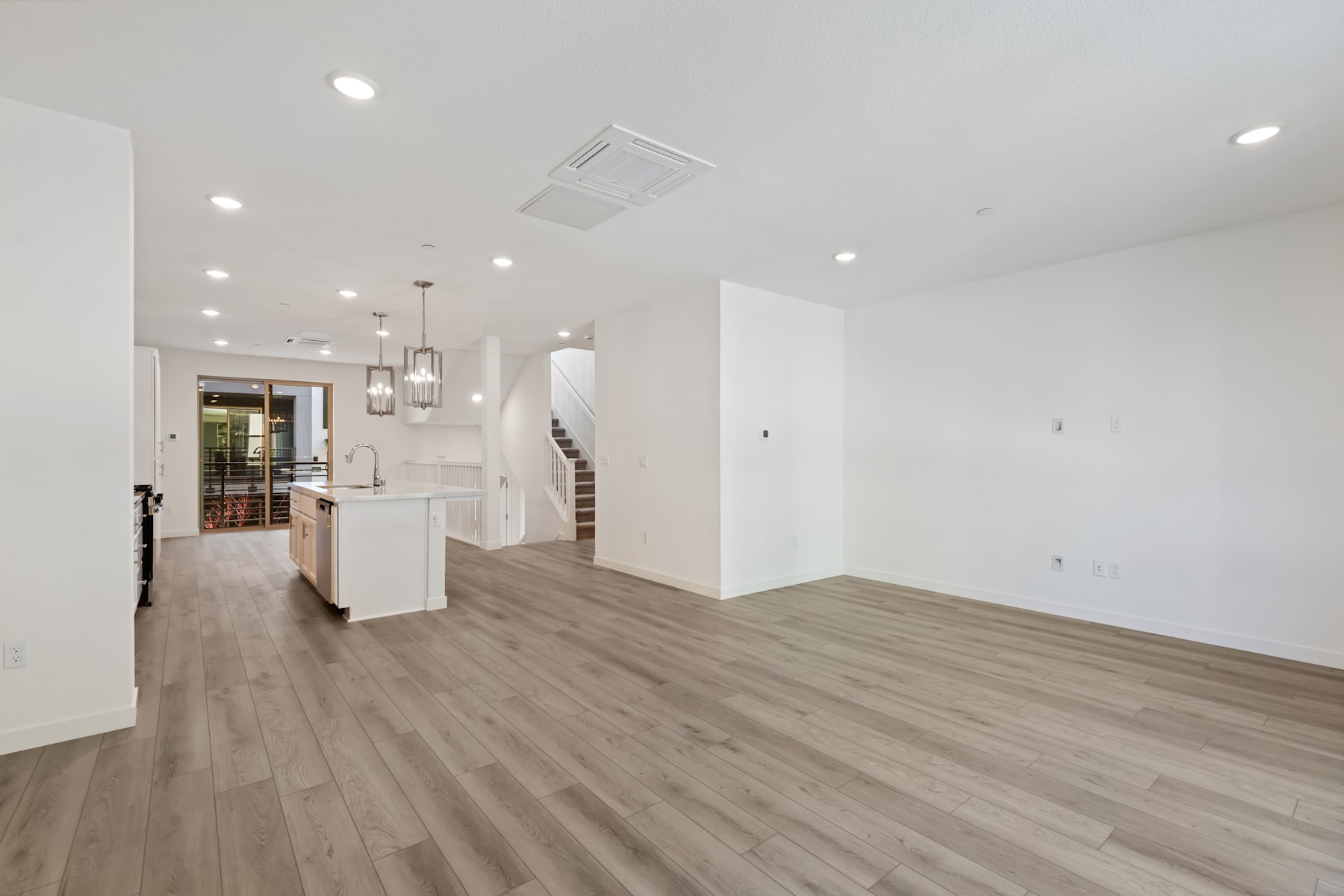 A spacious, open-concept living area with white walls, hardwood flooring, and recessed lighting, leading into a kitchen area with white cabinetry and appliances.