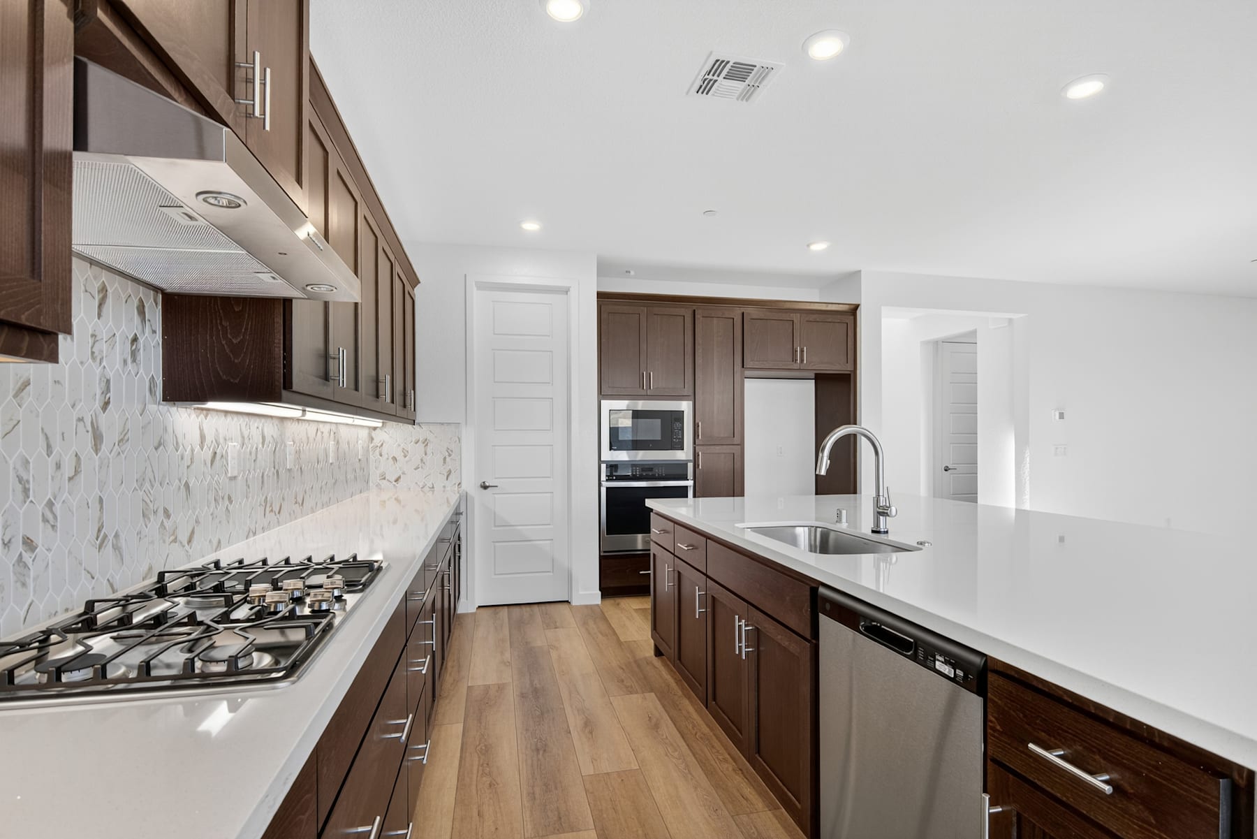 A modern, well-lit kitchen with dark wood cabinets, a gas stove, and a tiled backsplash, leading into a bright, open space.