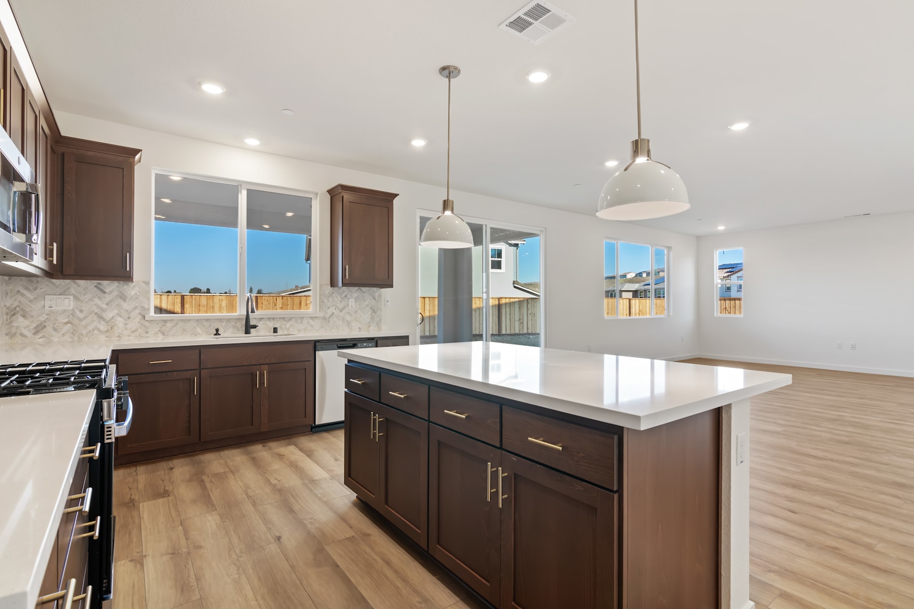 A modern and spacious kitchen with dark wood cabinets, a white countertop, and pendant lighting fixtures, set against a backdrop of large windows offering a view of the outdoors.