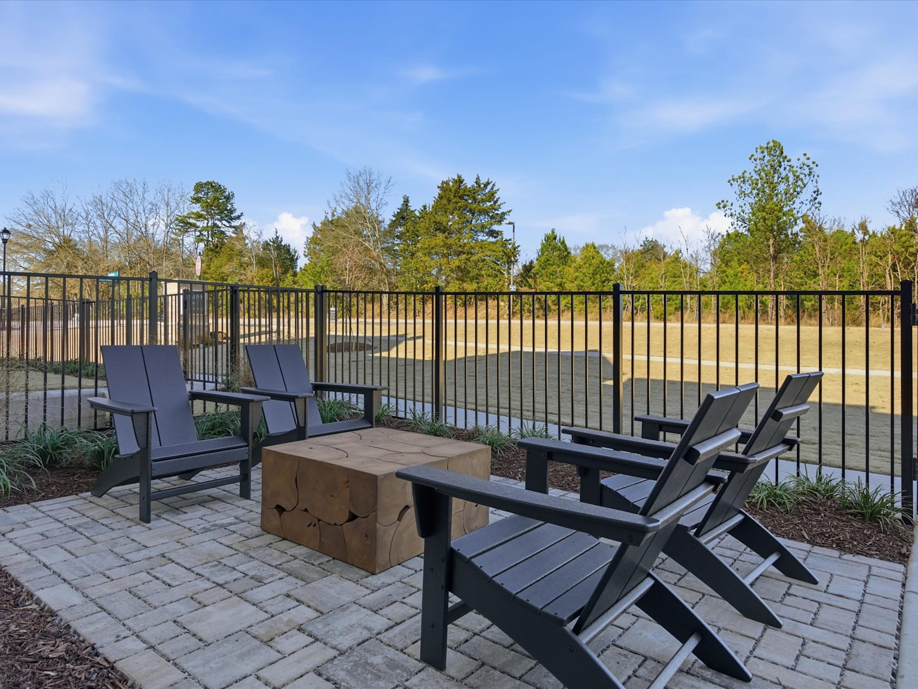 A paved patio area with black outdoor furniture, surrounded by a metal fence and a wooded landscape in the background.
