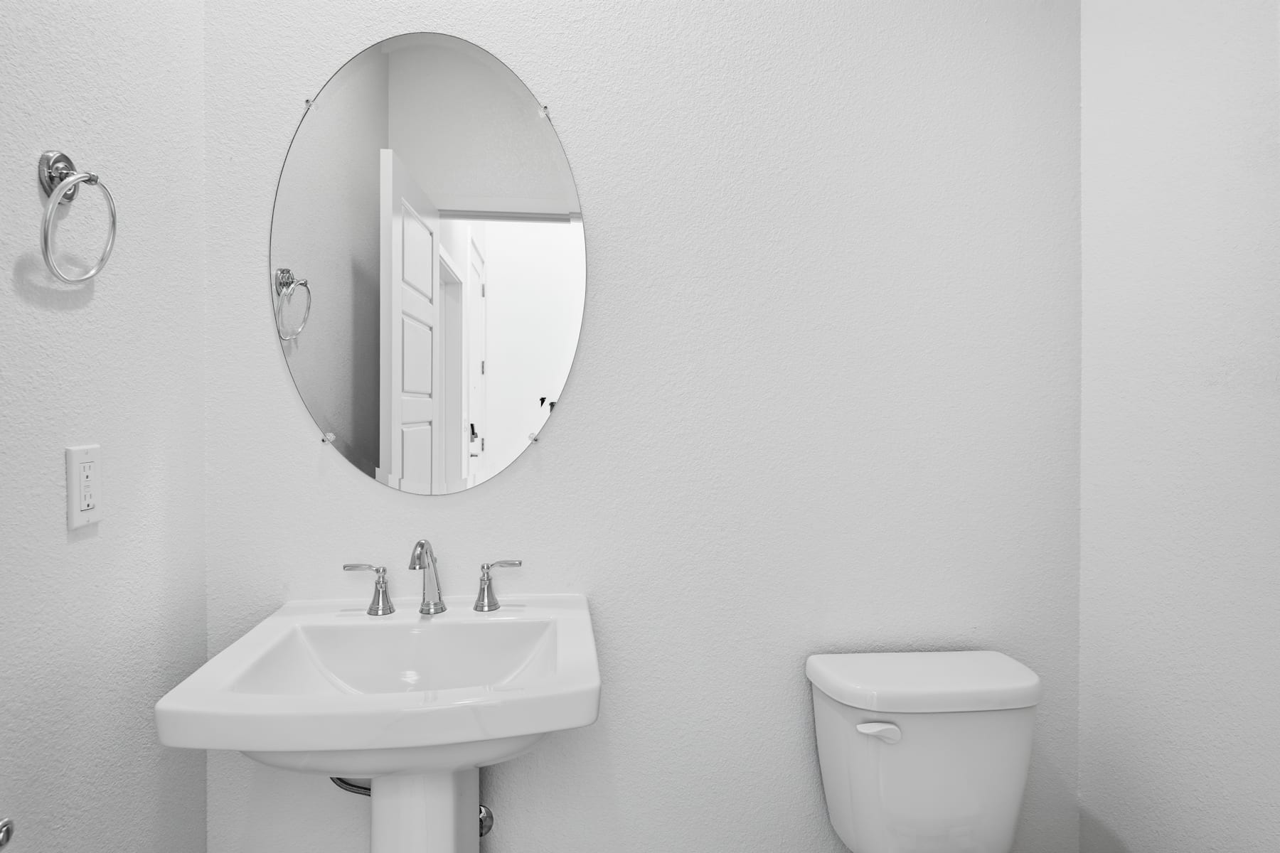 A minimalist bathroom with a white pedestal sink, an oval mirror, and a toilet against a plain white wall.