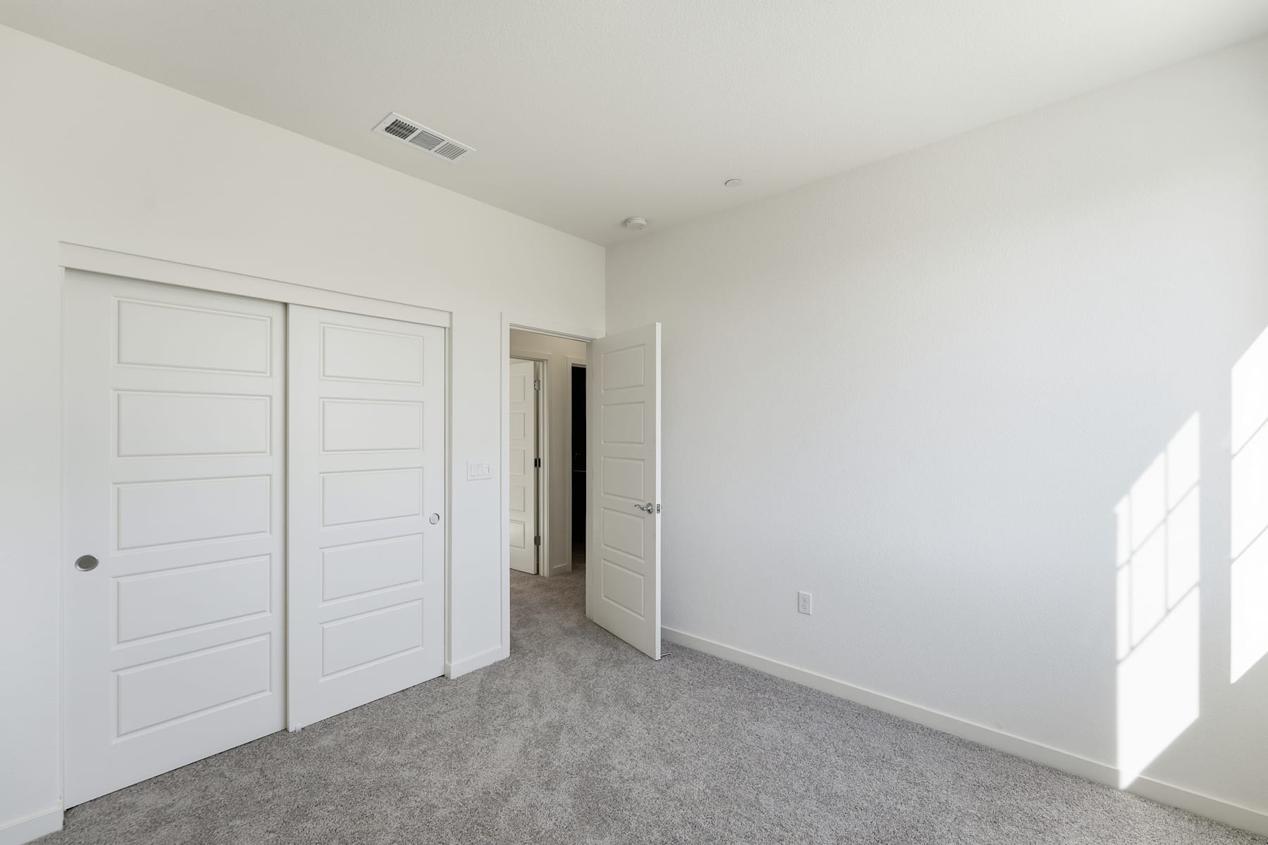 A spacious, minimalist bedroom with white walls, gray carpeting, and two white doors leading to other rooms.