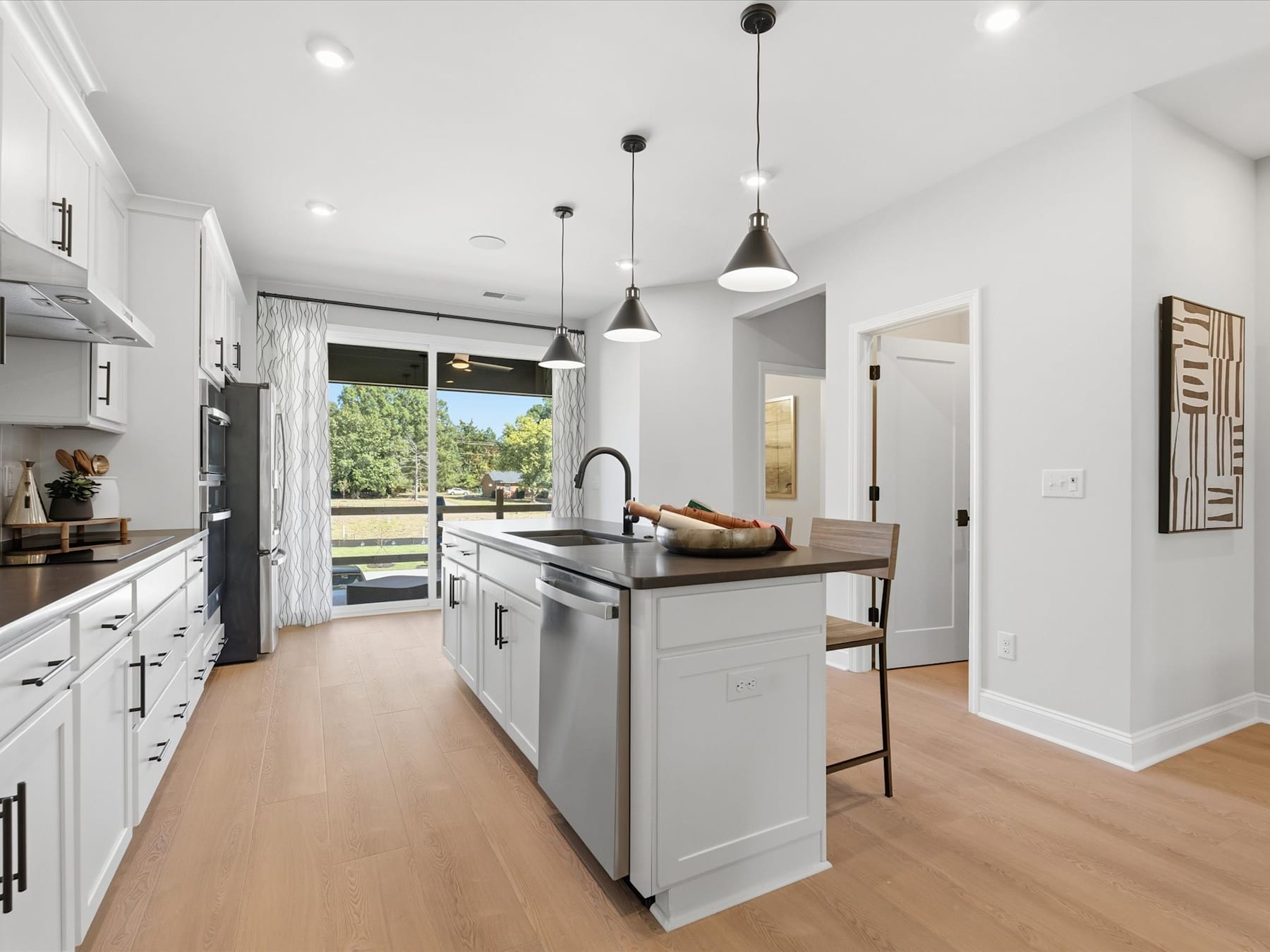 A modern, open-concept kitchen with white cabinets, stainless steel appliances, and a wooden island countertop, overlooking a scenic outdoor view through a sliding glass door.