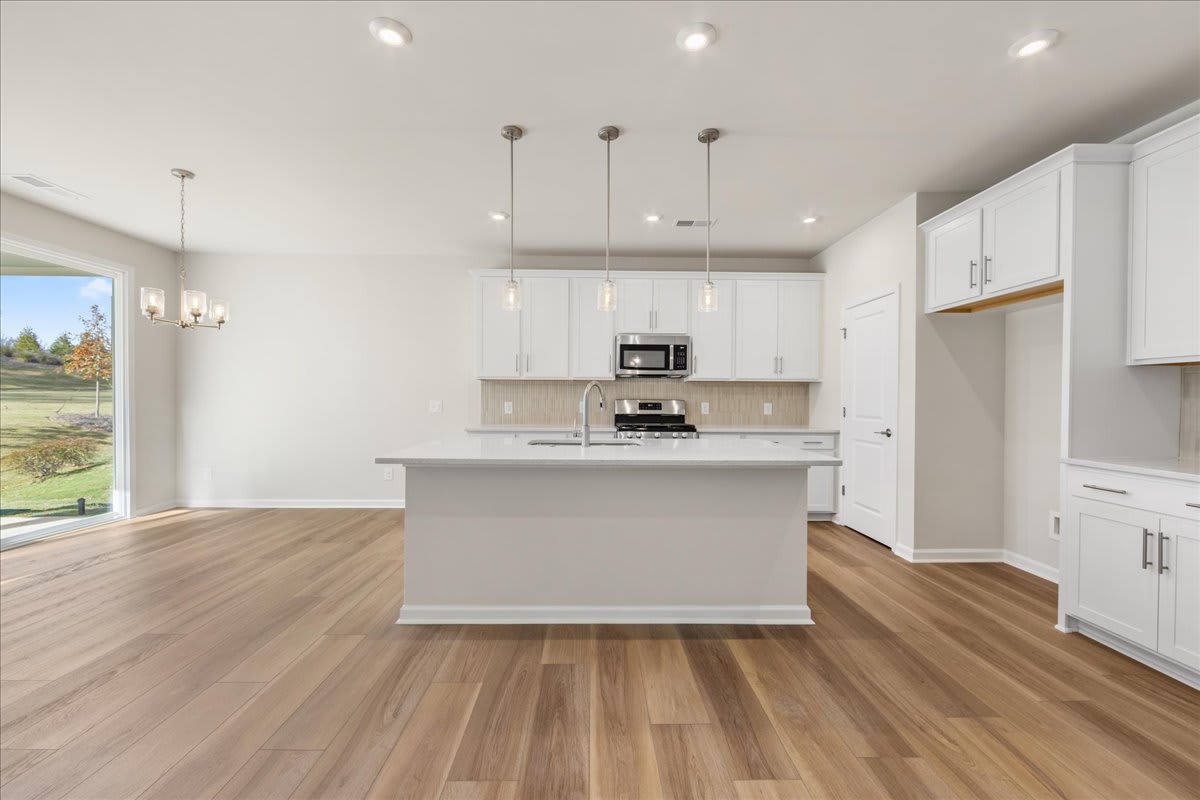 A modern, open-concept kitchen with white cabinets, a central island, and hardwood flooring, set against a backdrop of a scenic outdoor view through a large window.