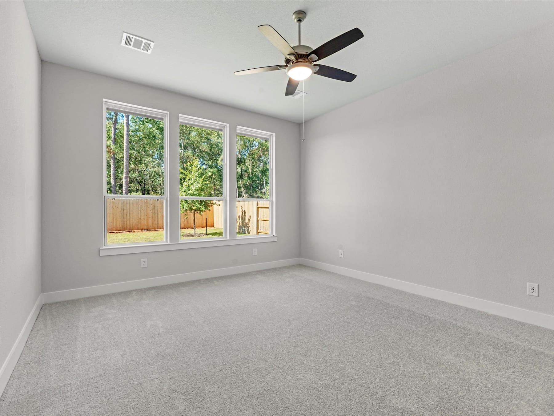 A spacious, well-lit room with a ceiling fan, large windows overlooking a backyard, and a neutral-colored carpet covering the floor.