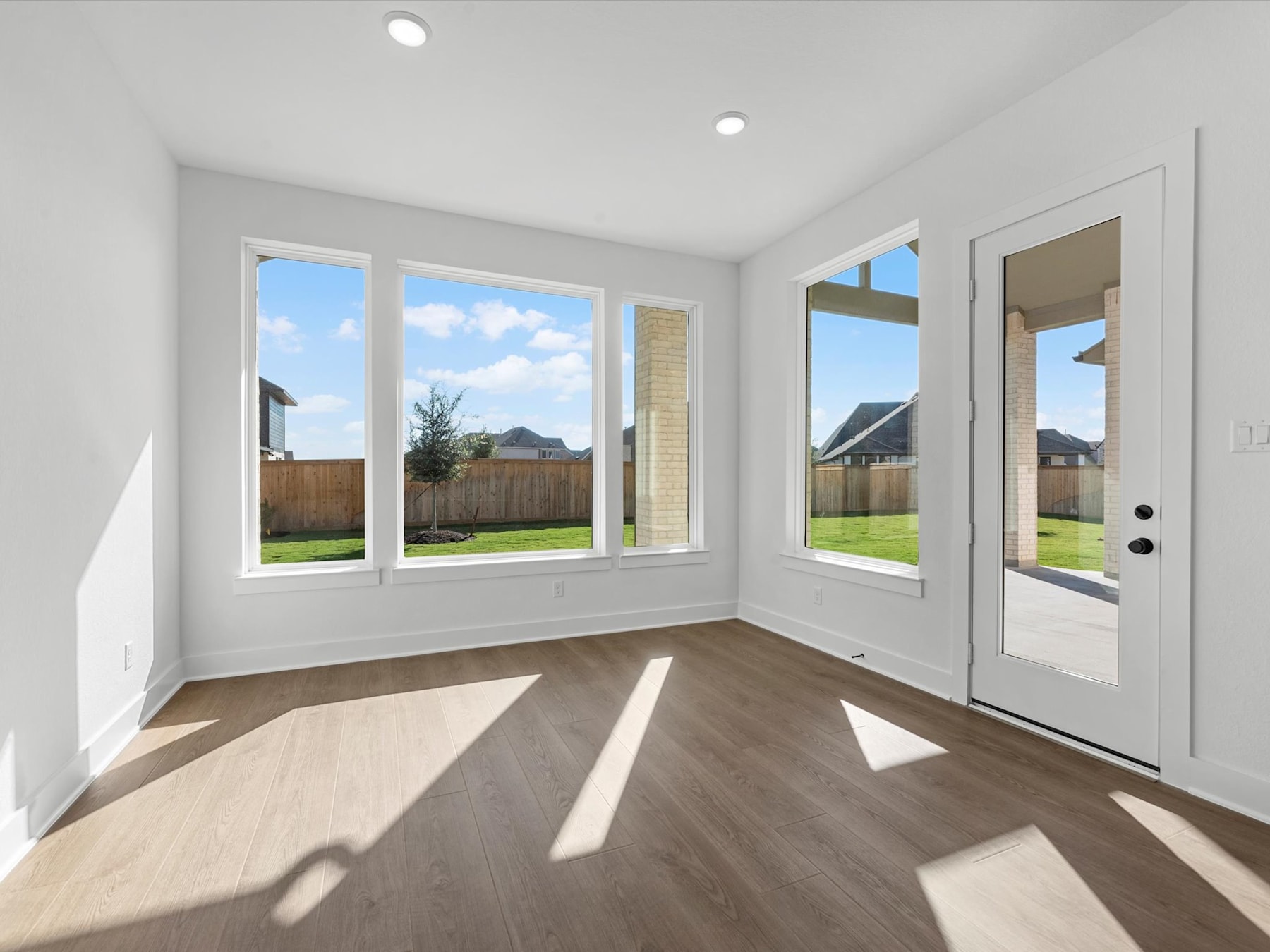 A bright and spacious room with large windows overlooking a grassy backyard, featuring a wooden floor and a white door leading outside.