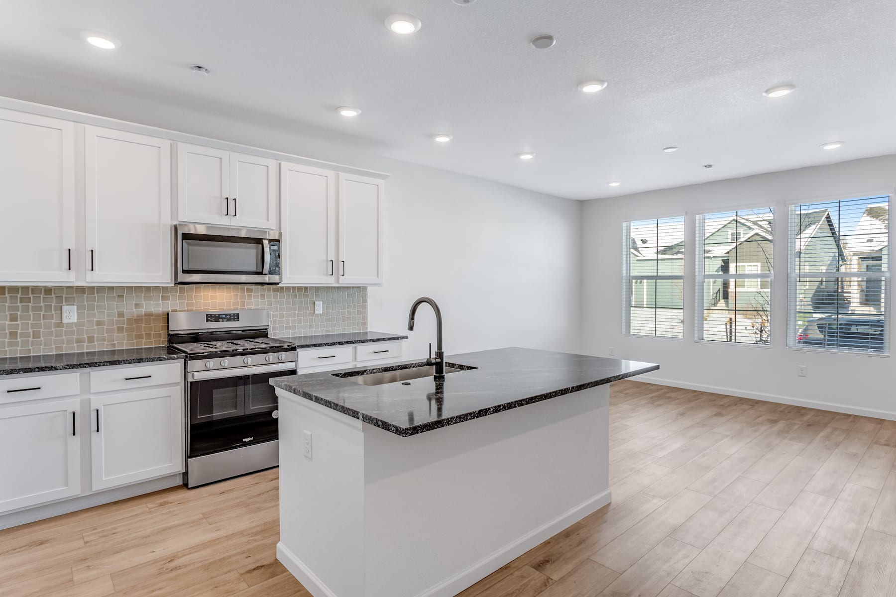 A modern, well-lit kitchen with white cabinets, black countertops, and stainless steel appliances, set against a backdrop of hardwood floors and large windows.