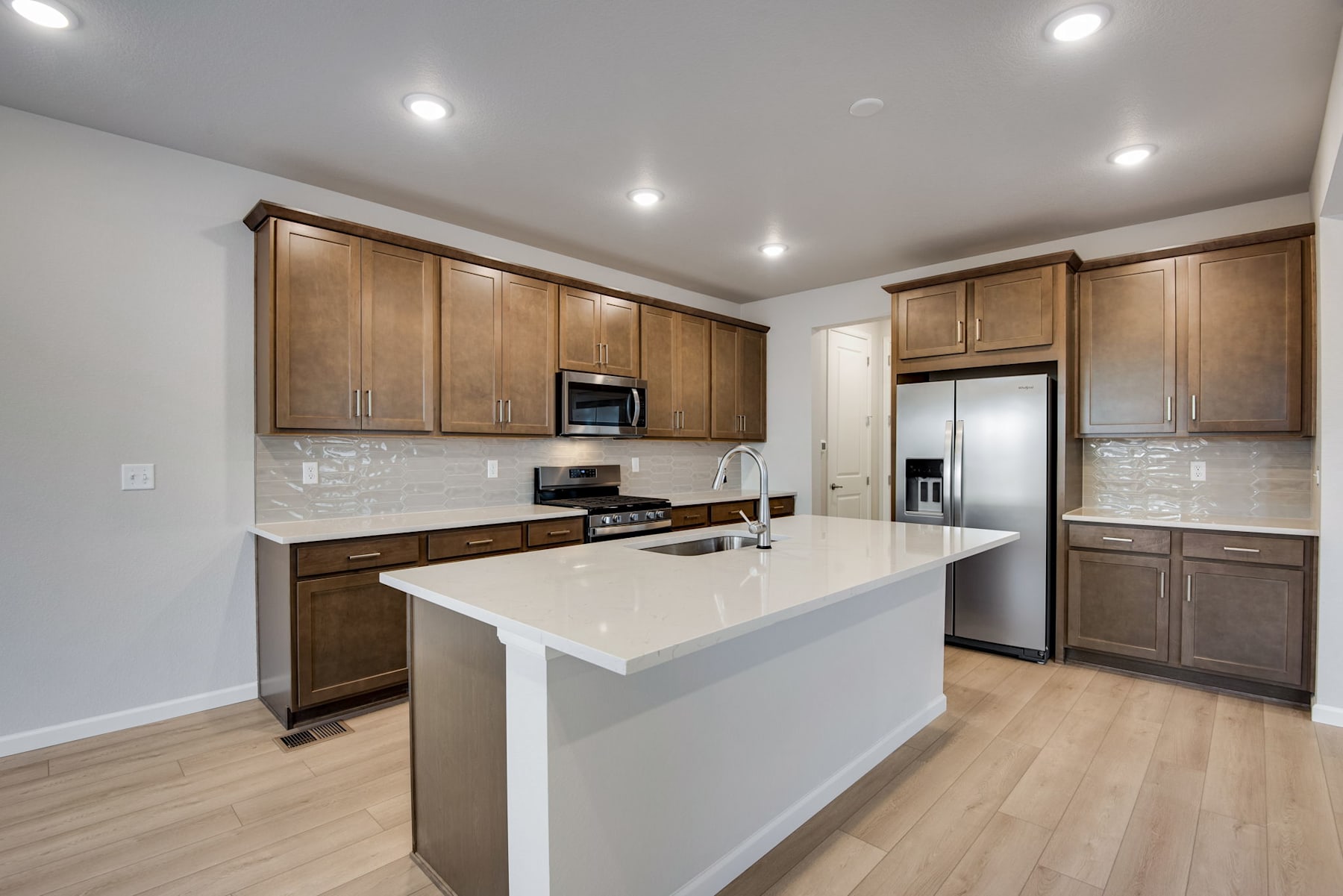 A modern, well-lit kitchen with wooden cabinets, a white countertop, and stainless steel appliances, set against a backdrop of light-colored hardwood floors.