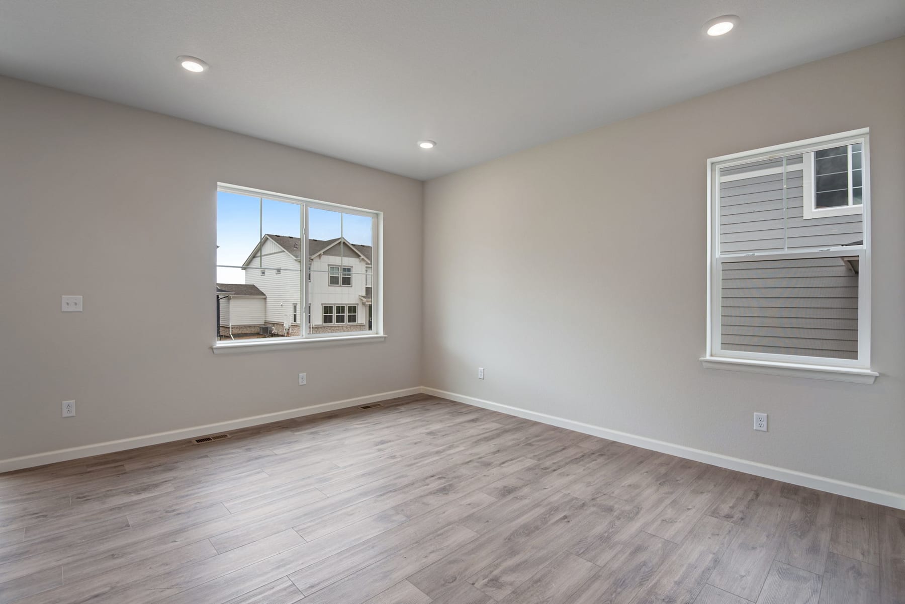A spacious, well-lit room with gray walls, hardwood floors, and a large window overlooking a residential neighborhood.