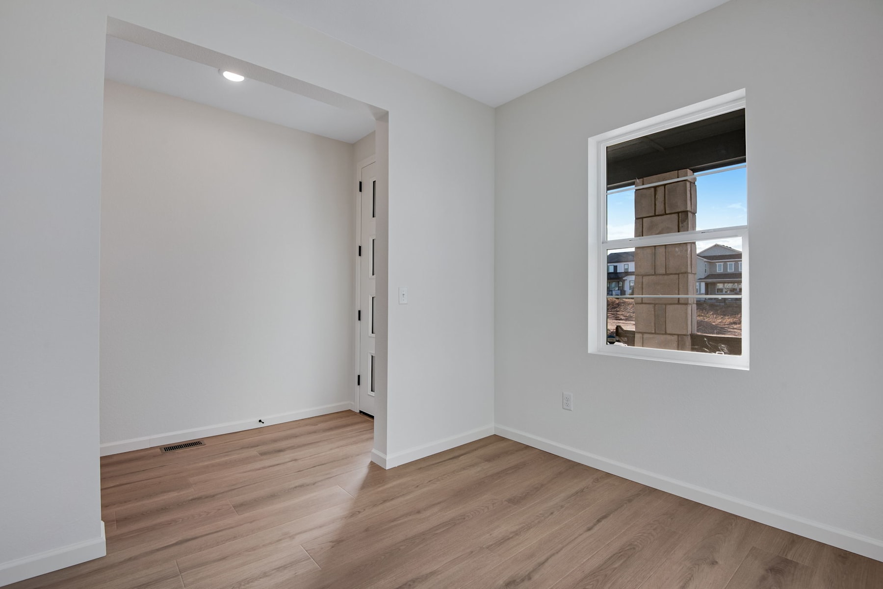 A spacious, well-lit room with white walls, a wooden floor, and a window overlooking a residential area.
