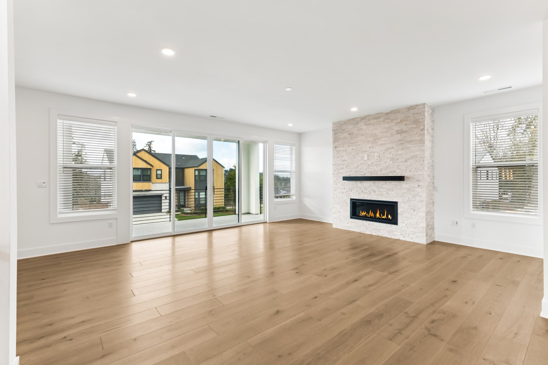 A spacious and bright living room with hardwood floors, a fireplace, and large windows overlooking a residential neighborhood.