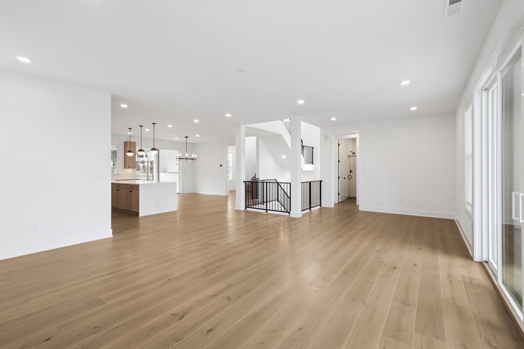 A spacious, open-concept living area with hardwood floors, white walls, and recessed lighting, leading into a kitchen area with wooden cabinetry.